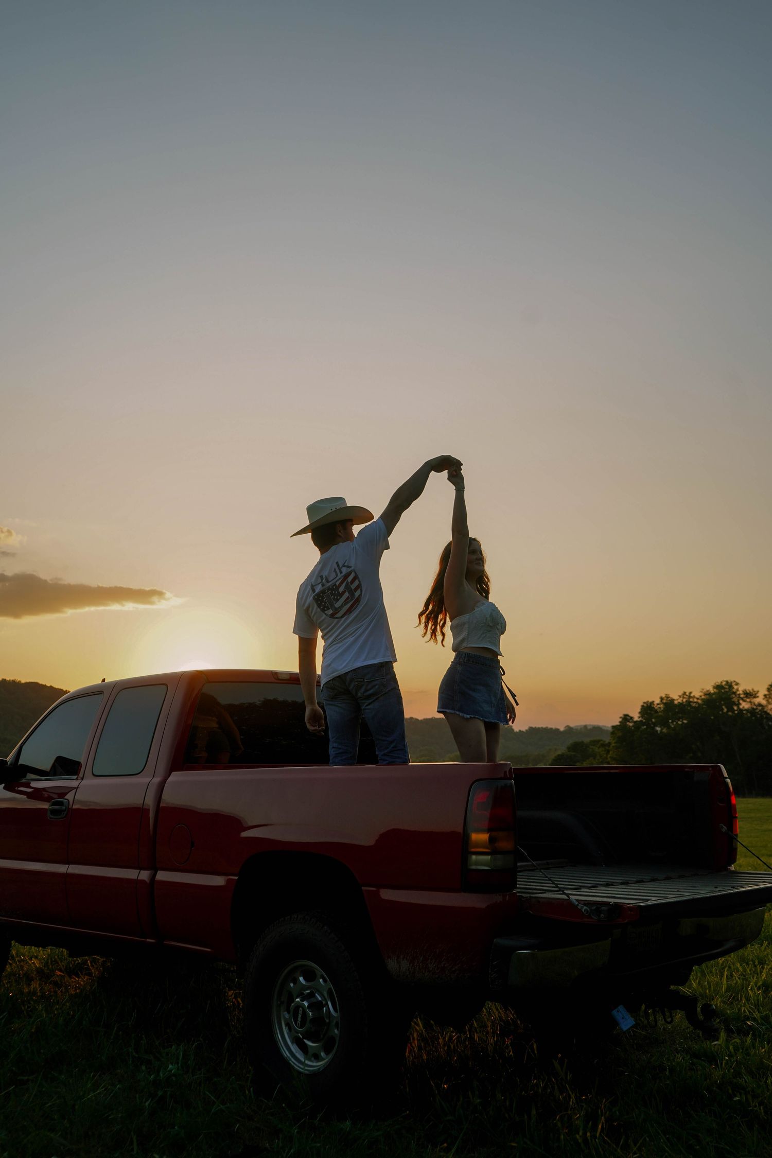 Couple dancing on the bed of a red pickup truck at golden hour sunset in a rural field.