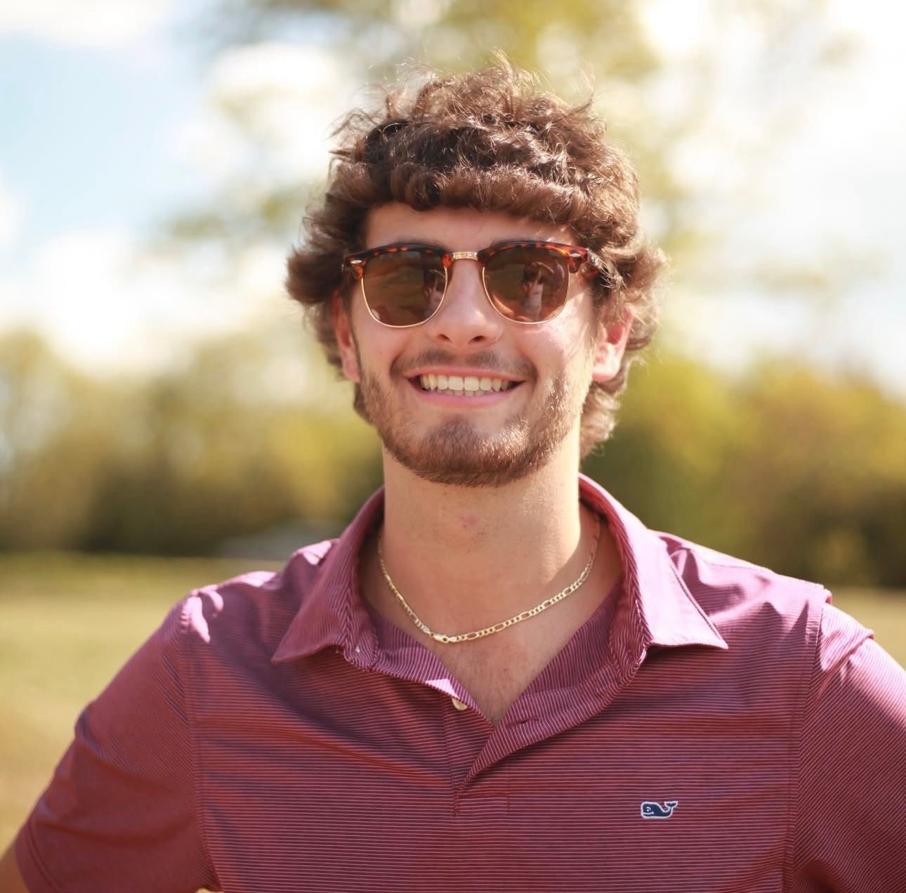 Smiling young man with curly hair wearing sunglasses and a pink Vineyard Vines shirt outdoors.