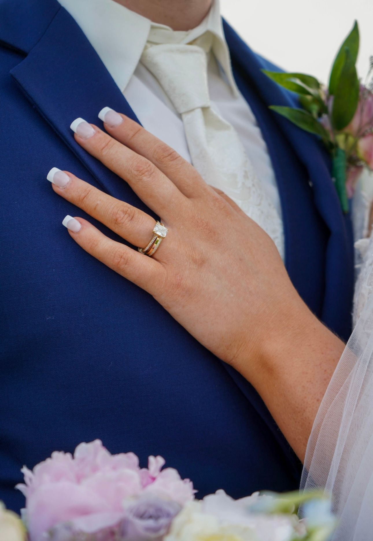 Close-up of bride's hand with gold engagement ring resting on groom's blue suit jacket.