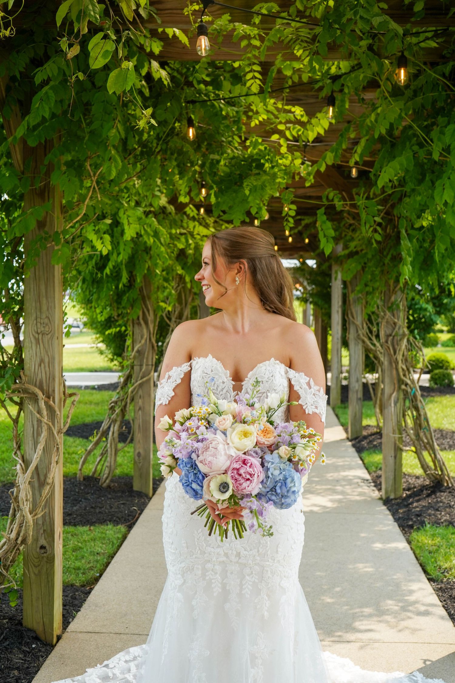 Smiling bride in lace mermaid gown holds large pastel floral bouquet under a green vine-covered pergola arch.
