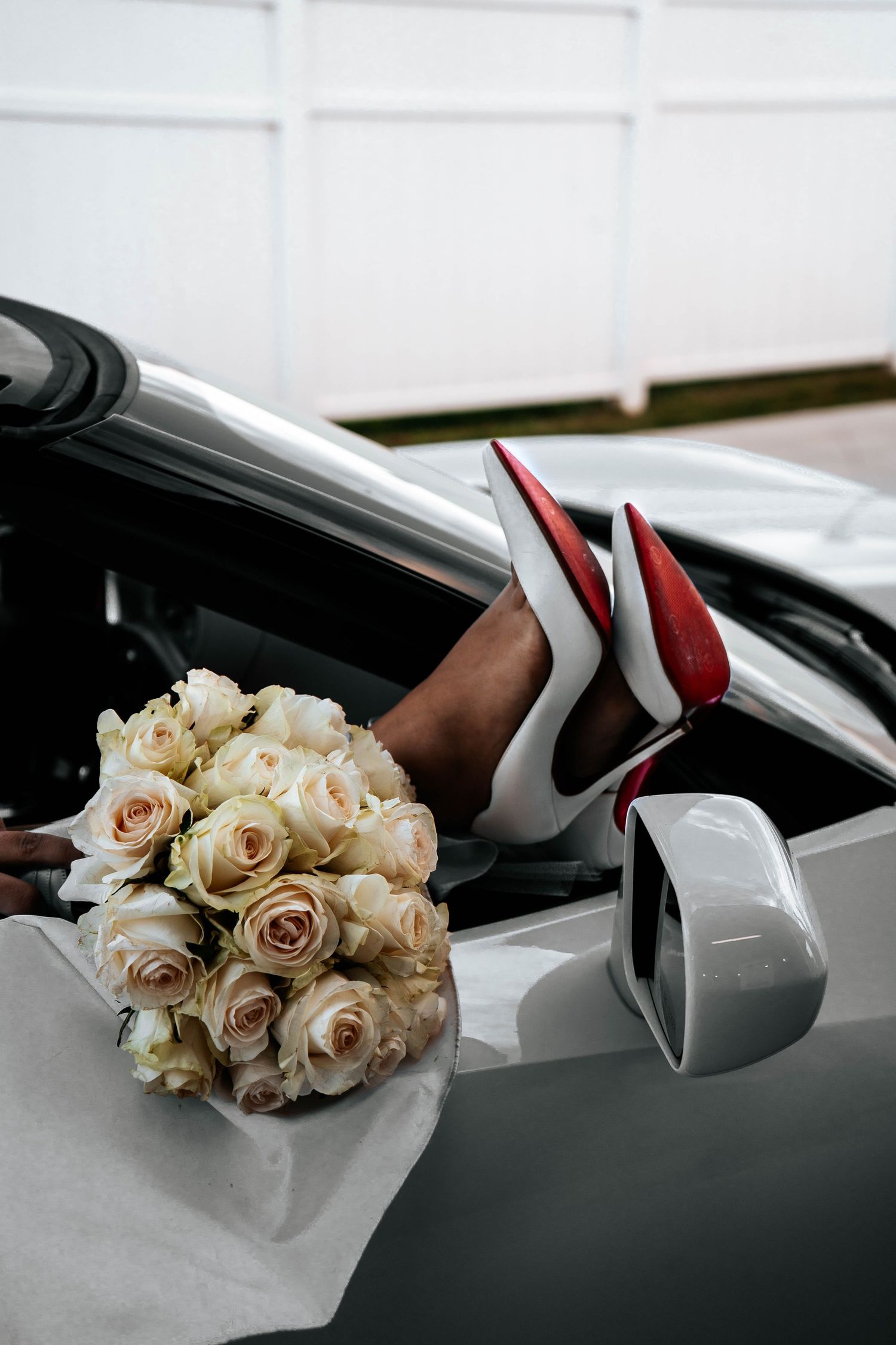 Bride's legs in red-soled heels dangle from white convertible car beside elegant white rose bouquet.