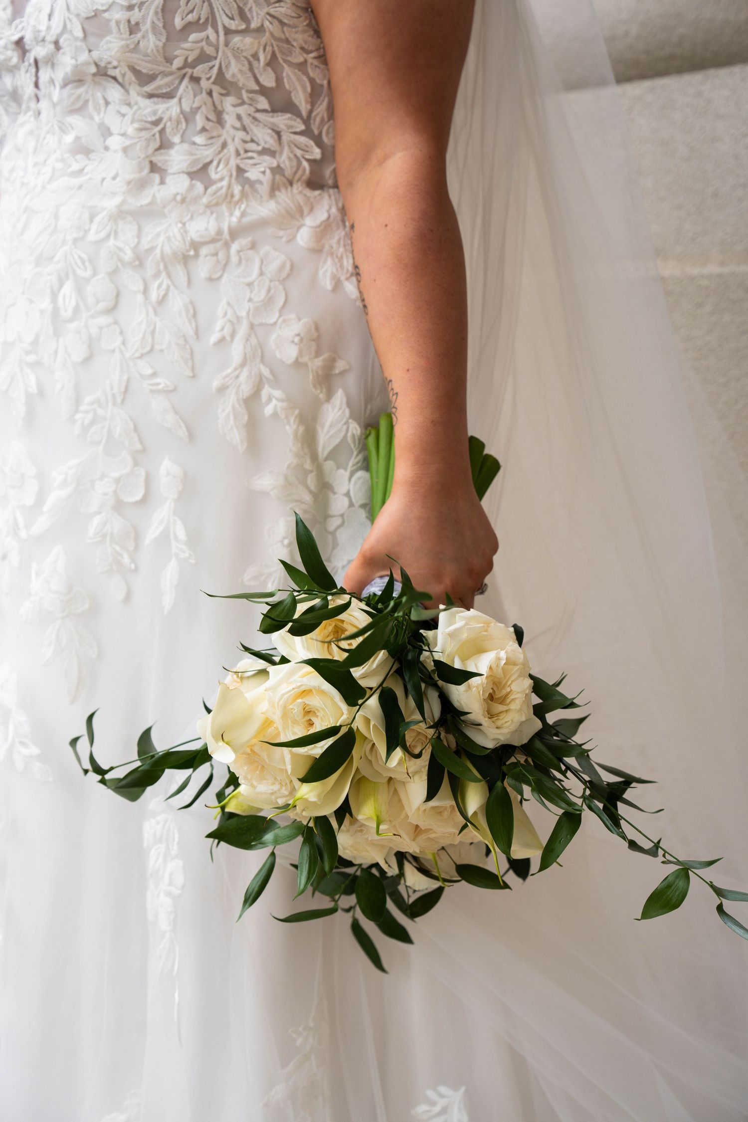 Bride in lace wedding gown holding elegant white rose and greenery bouquet at her side.