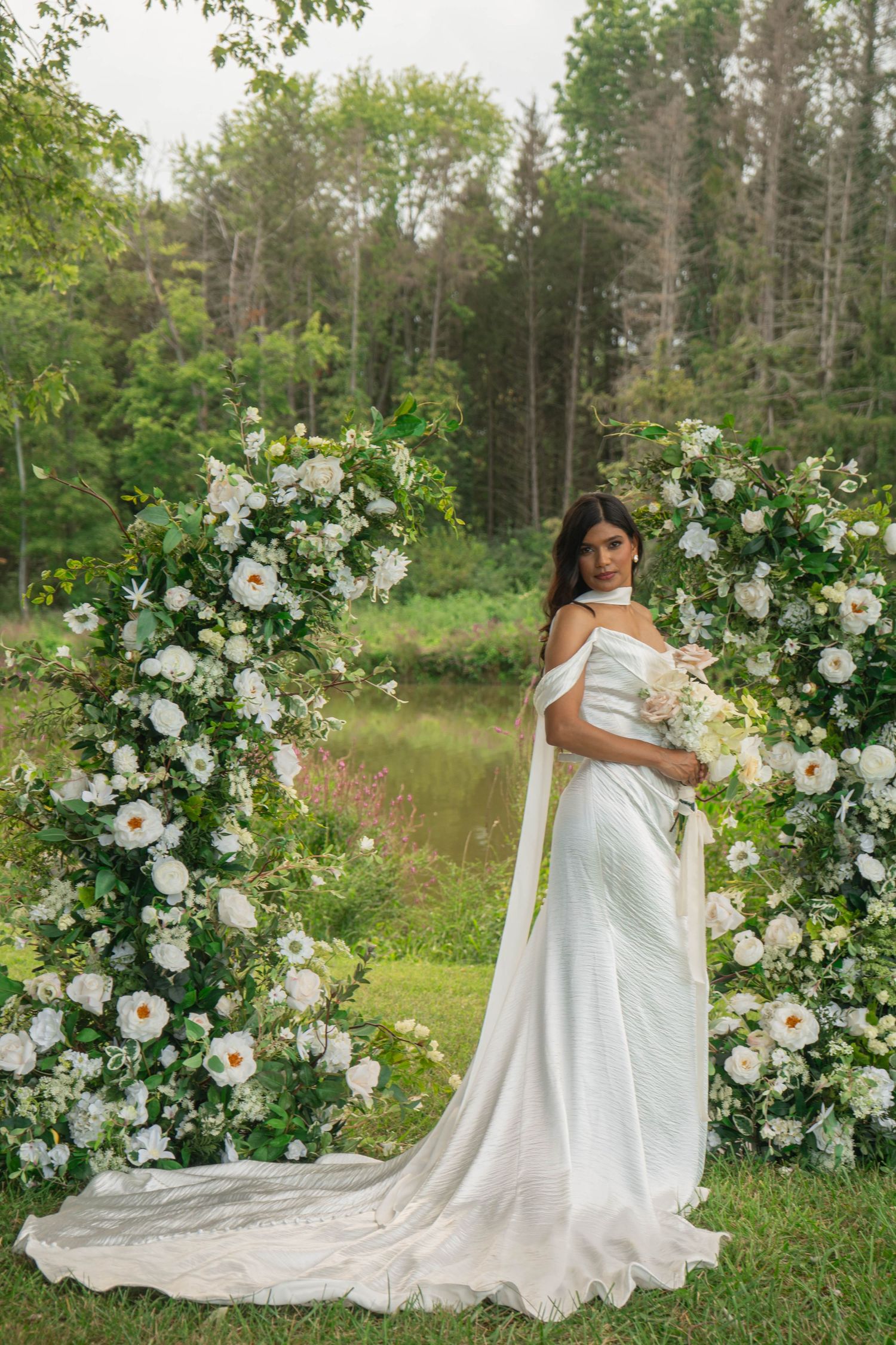Bride in white satin gown poses before a lush circular floral arch of white roses and greenery outdoors.