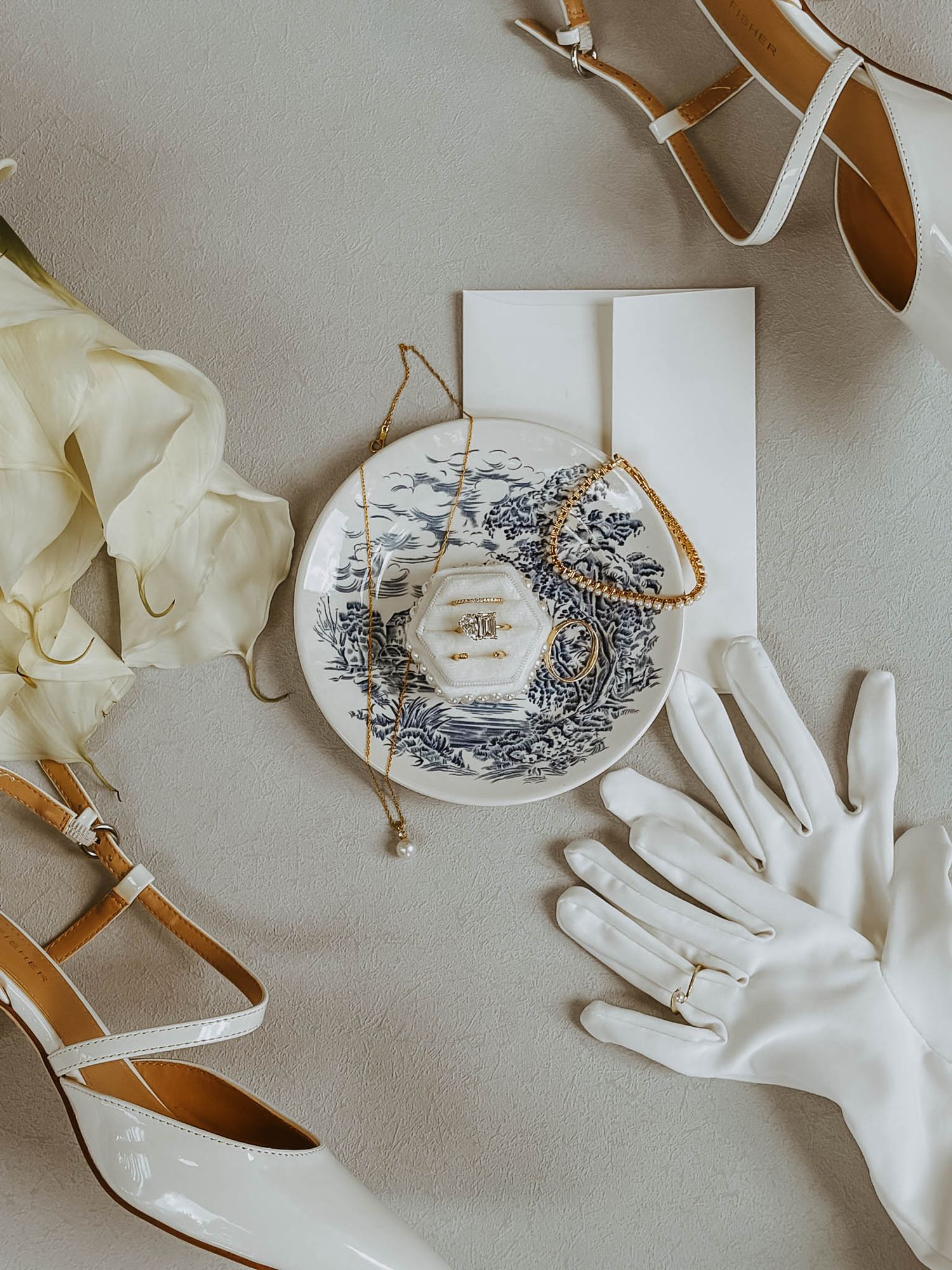 Bridal accessories flatlay featuring white heels, chinoiserie plate, gold jewelry, and white gloves.