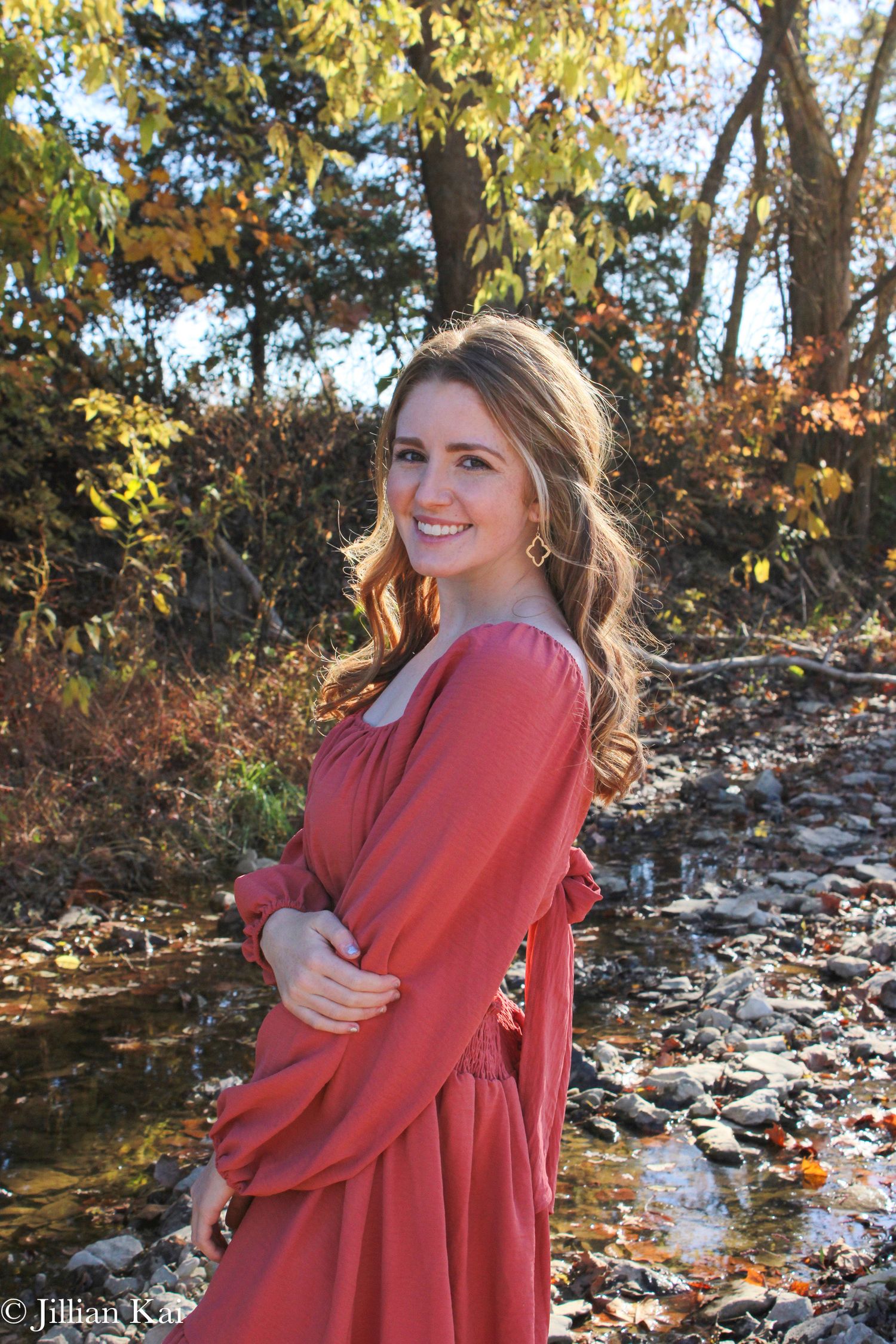 Young woman in coral dress smiling by rocky creek in autumn forest.