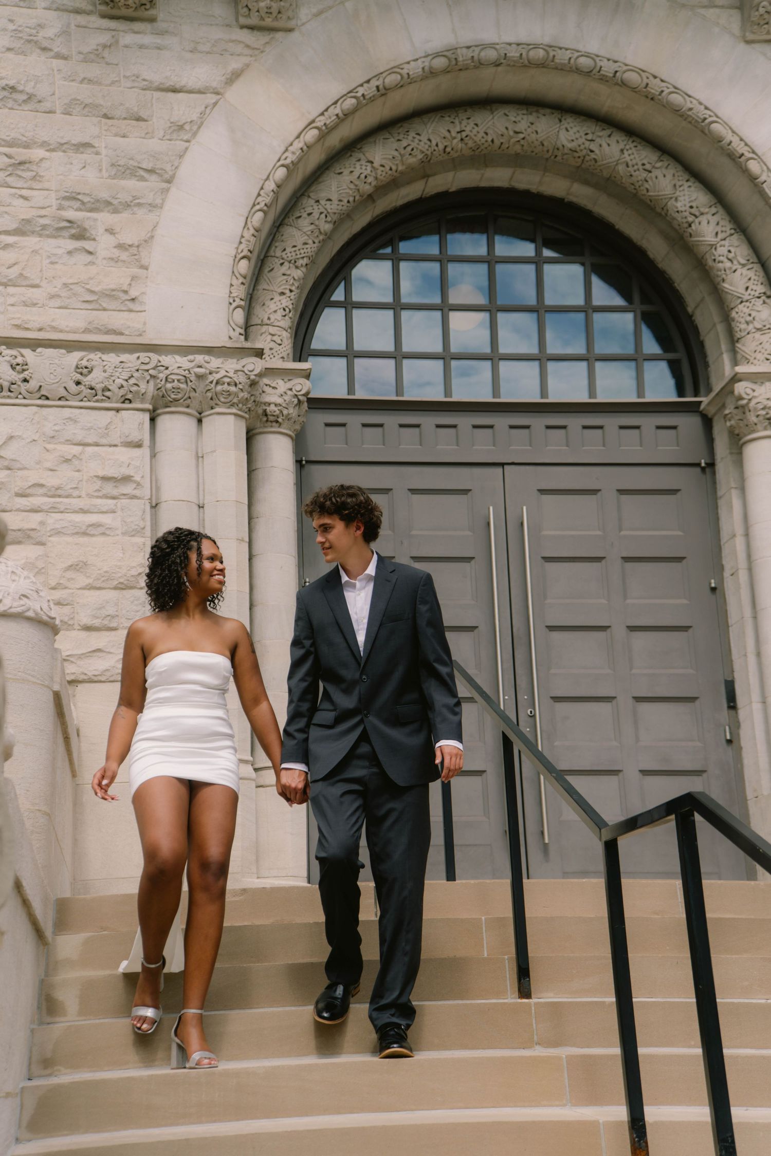 Smiling couple in wedding attire holding hands while descending stone steps of a classic building.