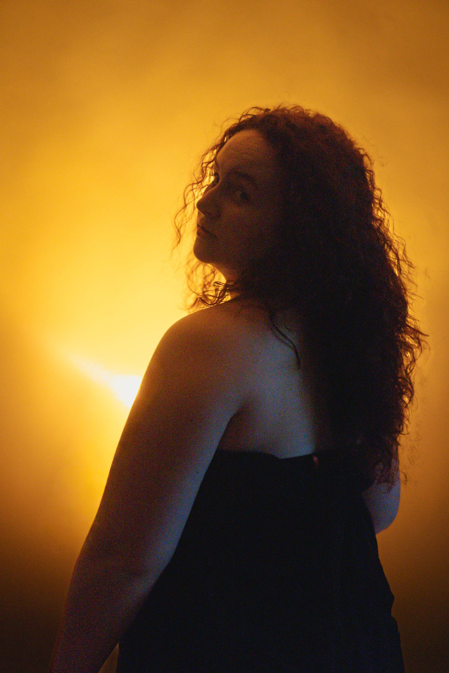 Silhouette of curly-haired woman in black top looking over shoulder against vibrant golden yellow background.