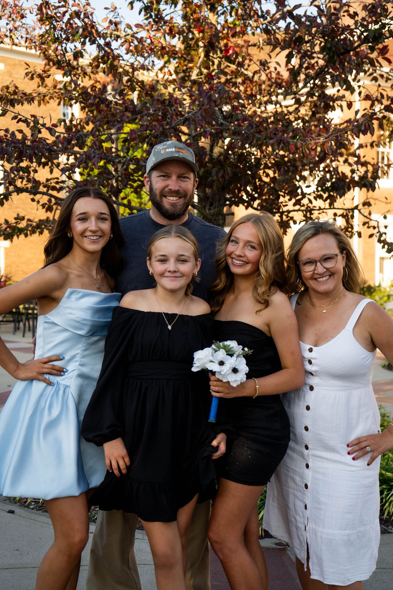 A family of five poses together outdoors under a purple-leafed tree, with three girls in formal dresses for prom.