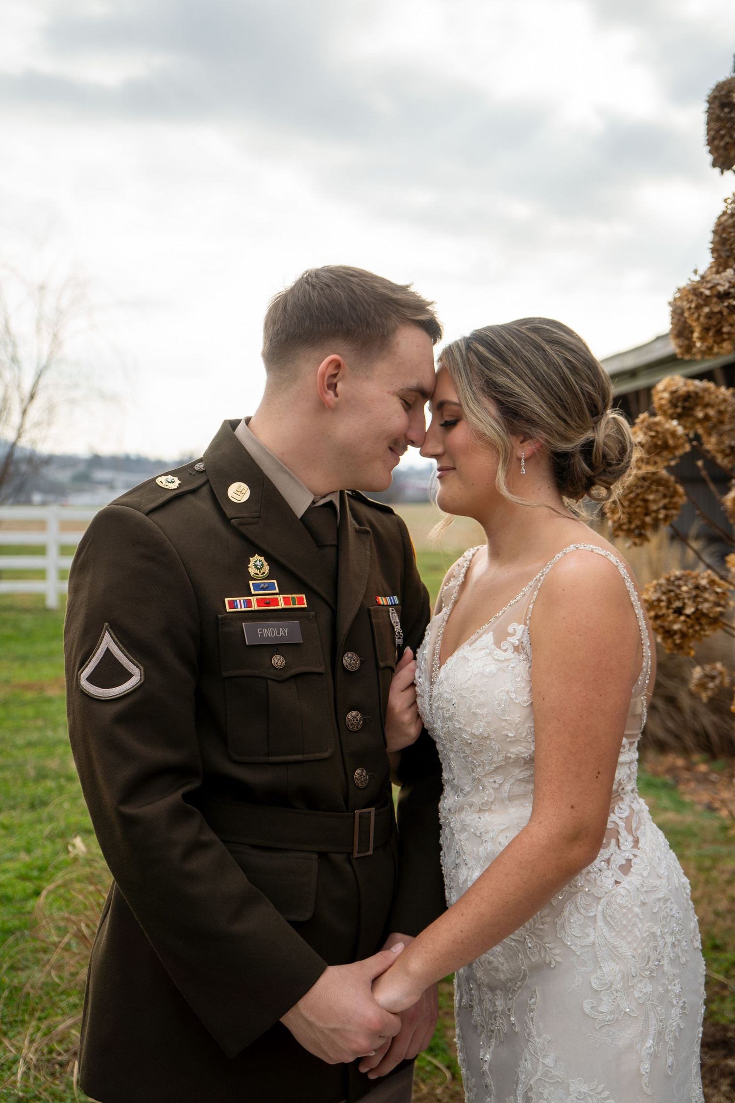 Military groom in dress uniform and bride in lace wedding gown touching foreheads outdoors.