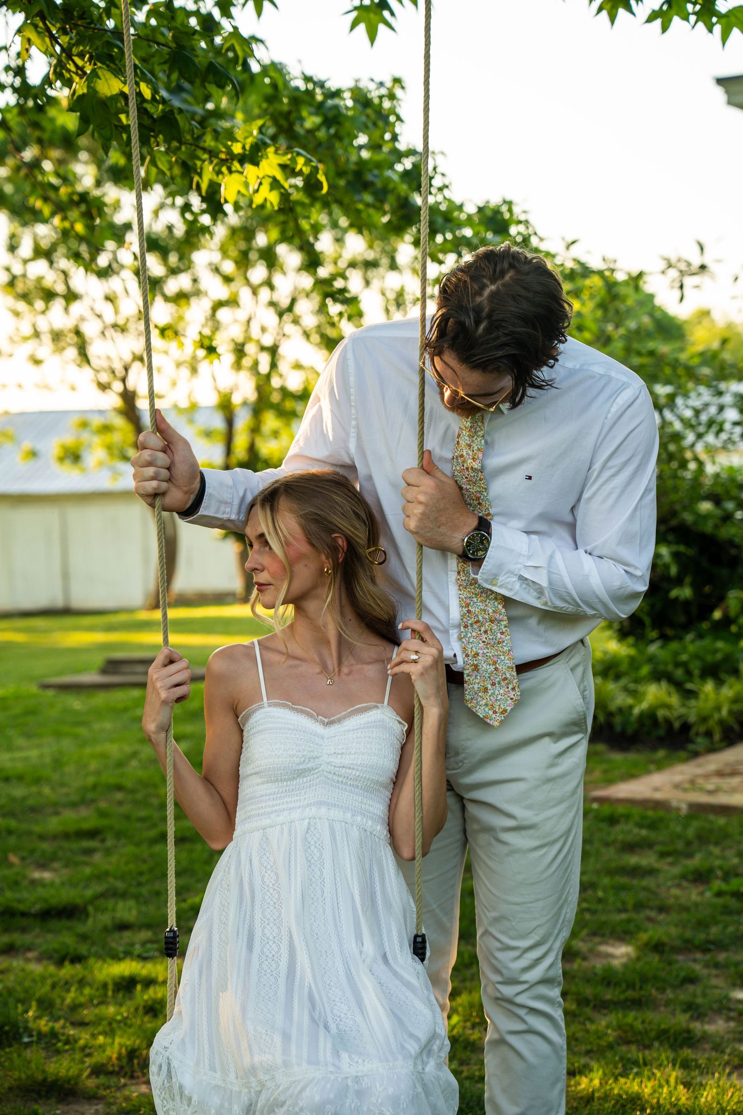 Bride in white dress sits on garden swing while groom in white shirt and floral tie stands behind her outdoors.