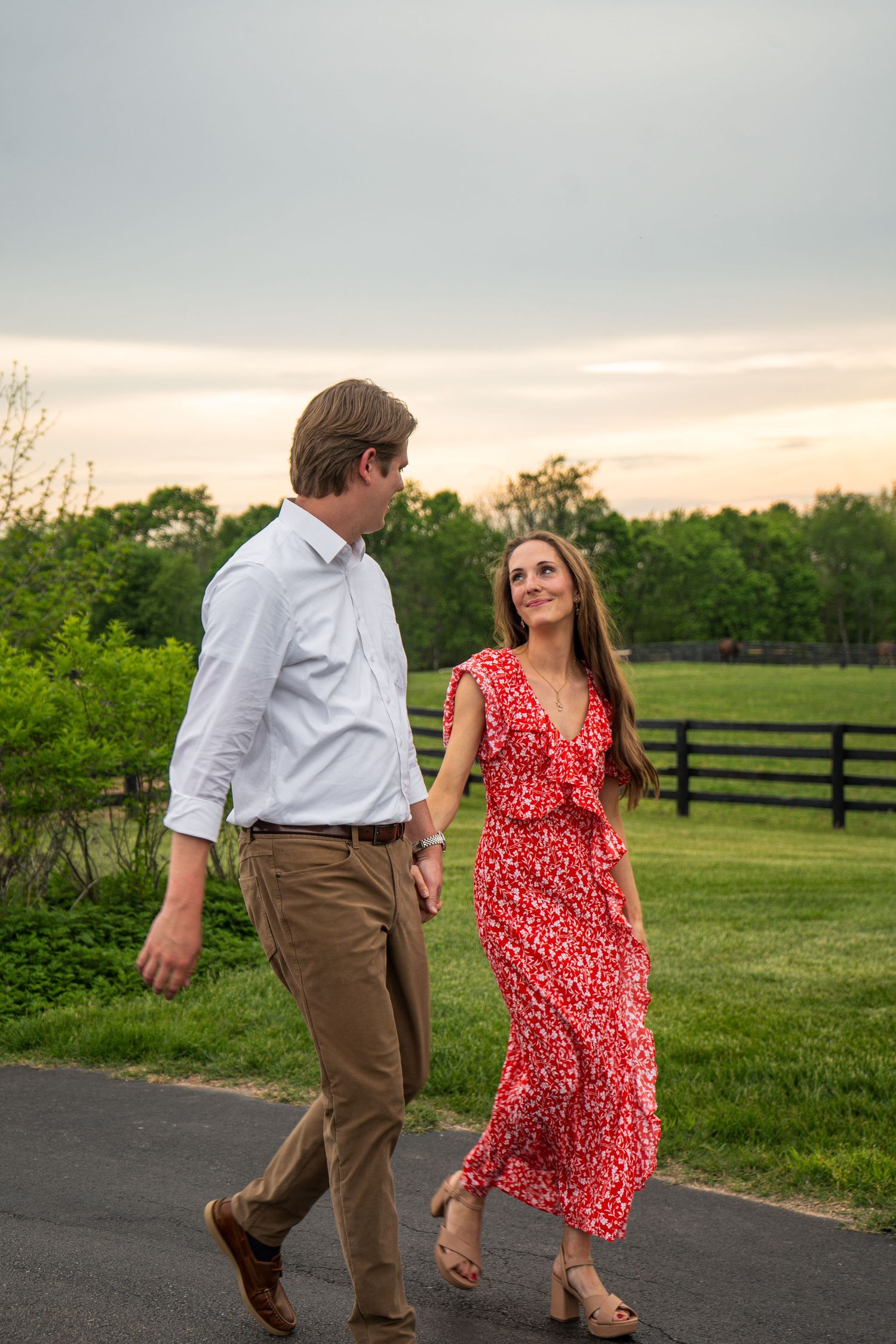 Engaged couple walking hand in hand along a path at sunset with green fields and fence behind.