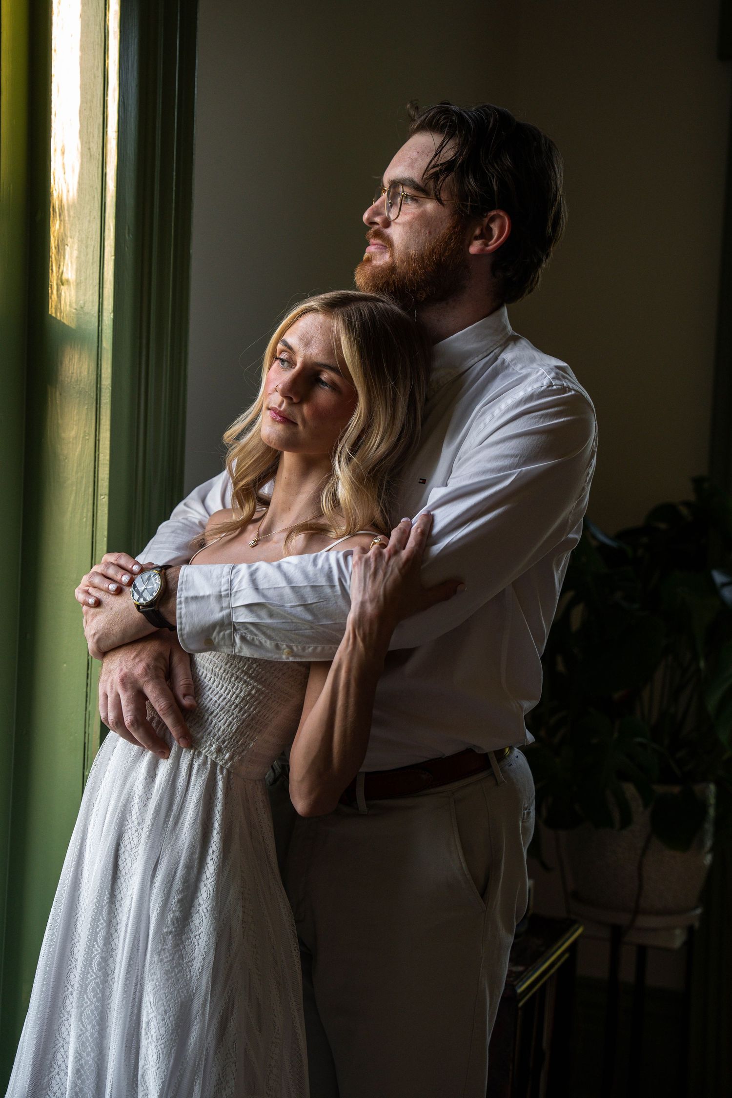 Couple embraces by a sunlit window, bride in white dress and groom in white shirt looking outward.