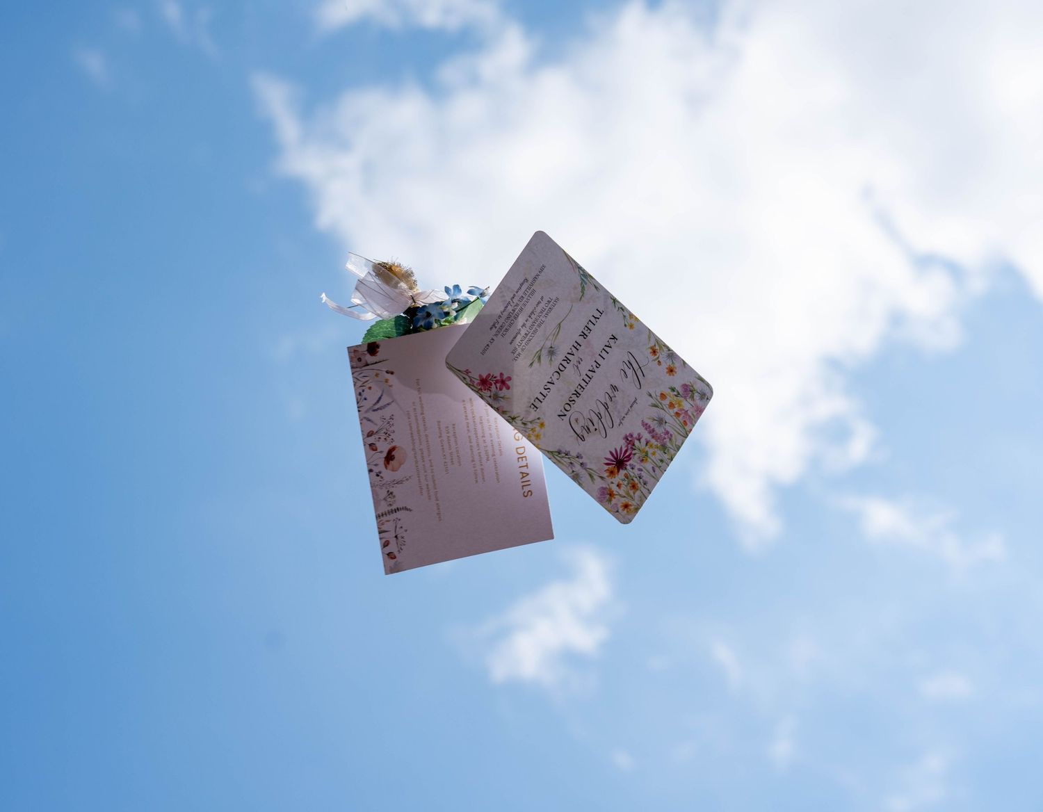 Floral wedding invitation cards tossed in the air against a bright blue sky with white clouds.