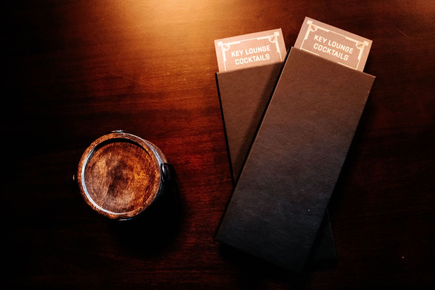 Two black menu holders with gold labels beside a glass of whiskey on a dark wooden table.