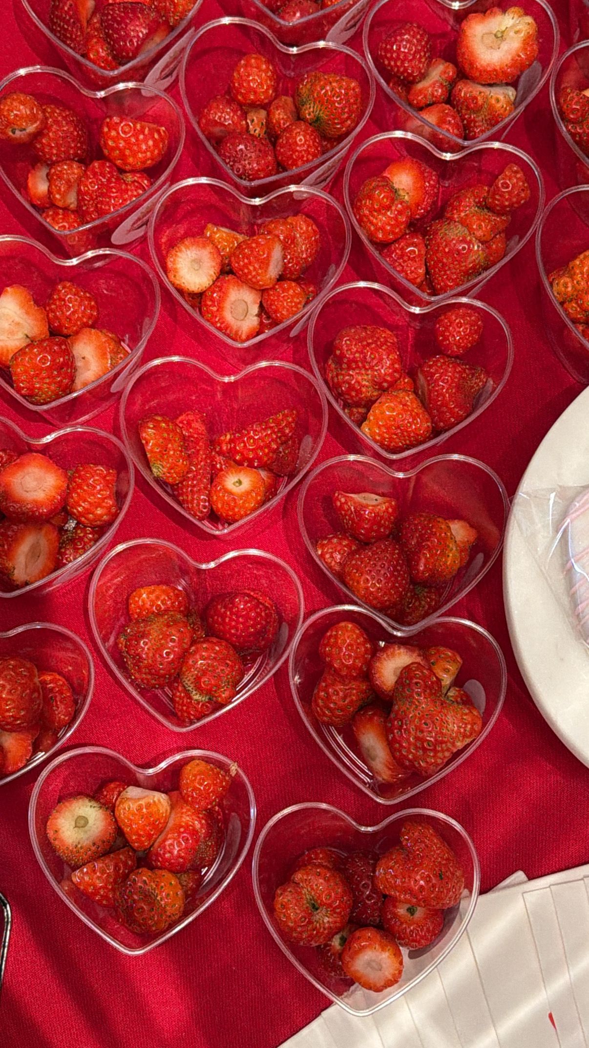 Heart-shaped clear bowls filled with fresh red strawberries arranged on a red tablecloth for a Valentine's Day event.