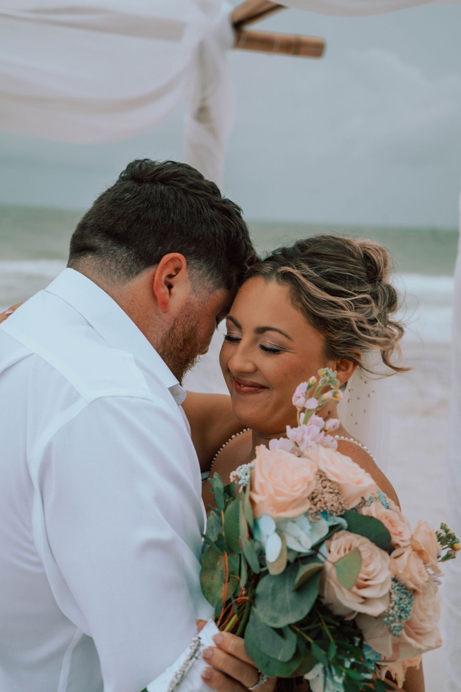 Bride smiling tenderly as groom nuzzles her cheek at a romantic beach wedding ceremony.
