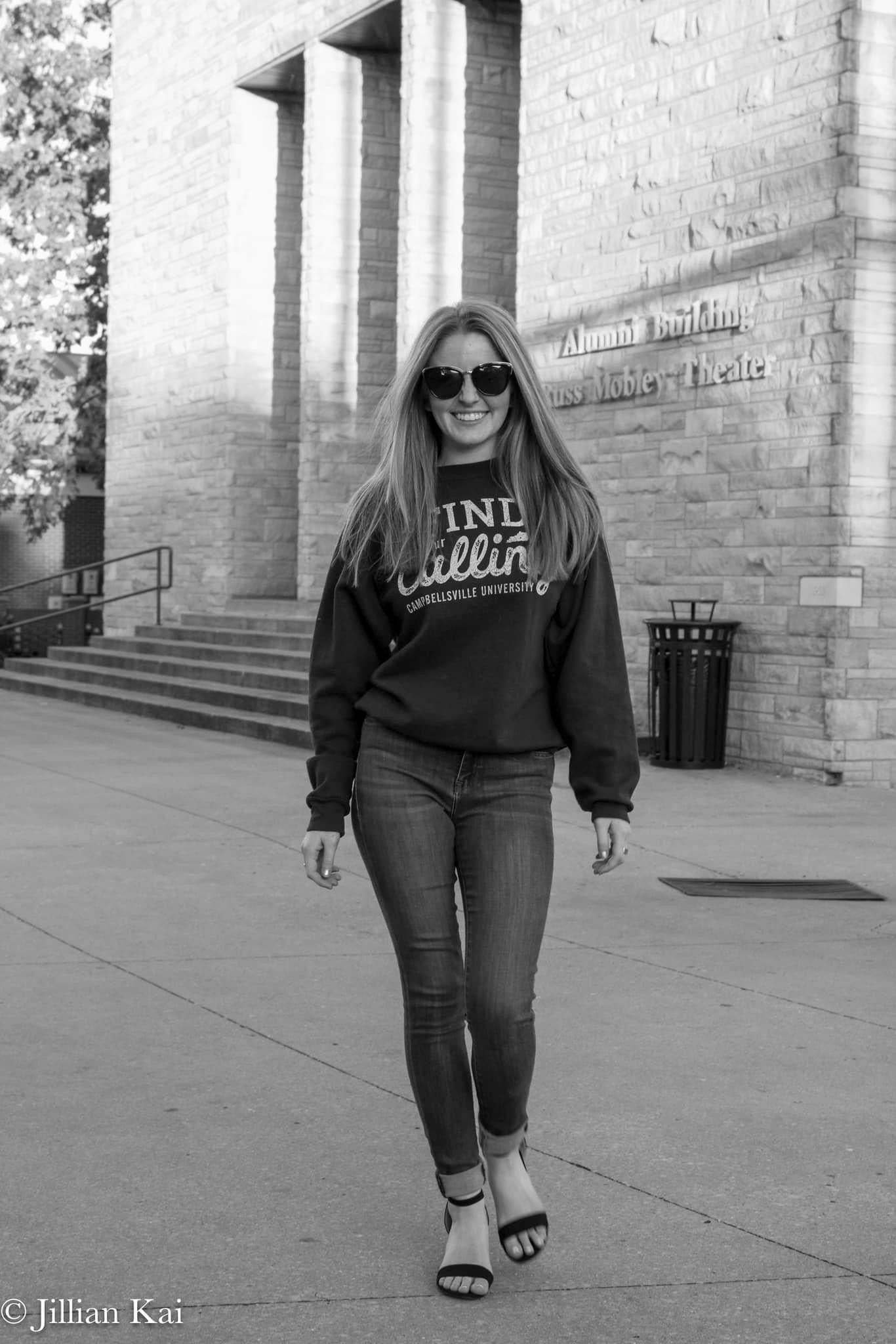 Blonde woman in Campbellsville University sweatshirt and sunglasses walking near Alumni Building in B&W.