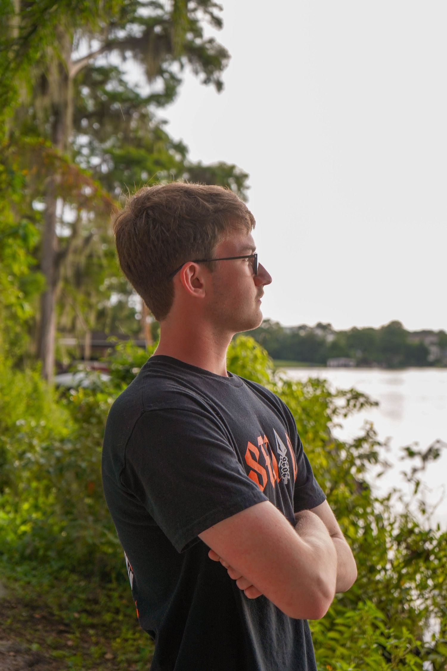 Young man in dark t-shirt with arms crossed looking at lake surrounded by trees.