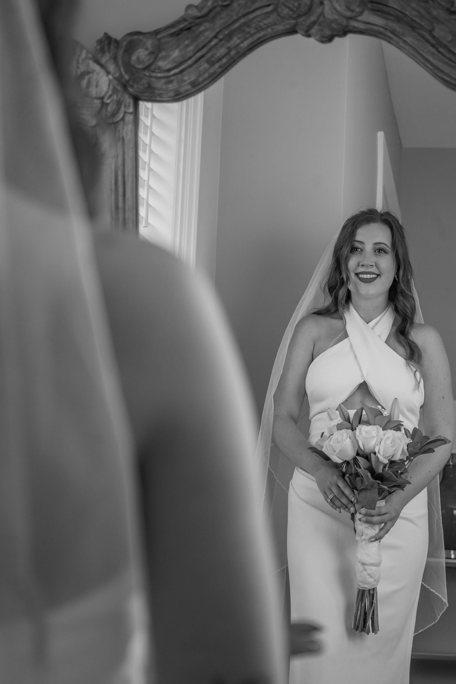 Black and white photo of smiling bride in halter wedding dress holding rose bouquet reflected in ornate mirror.