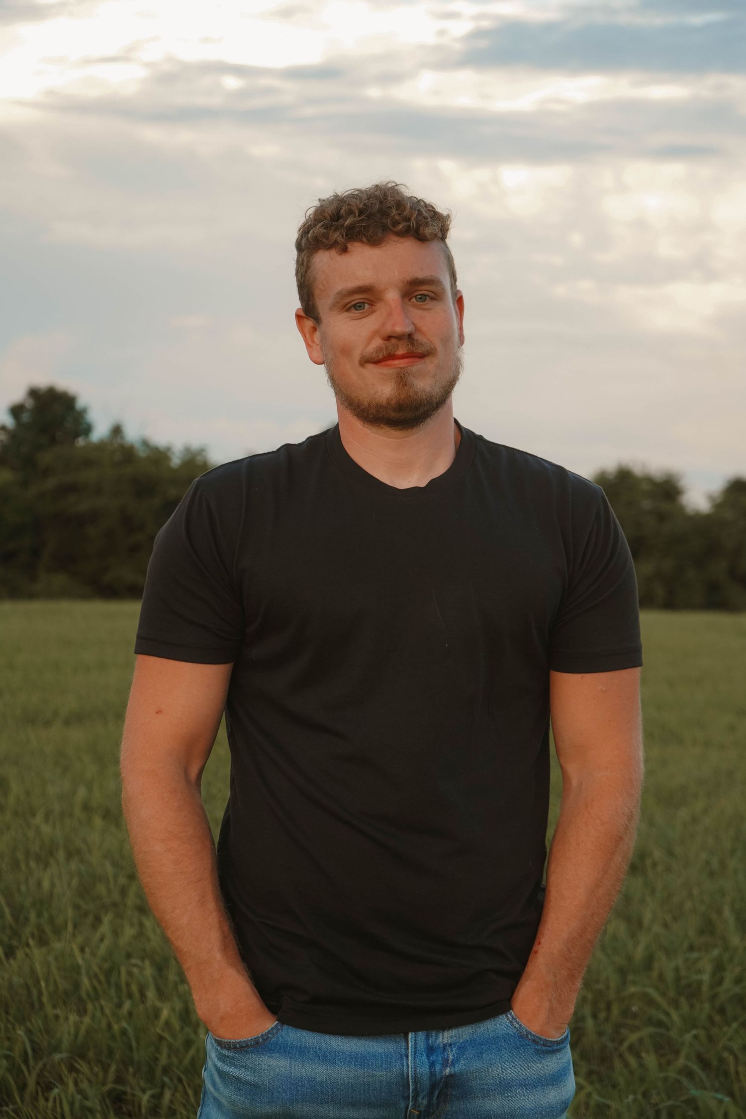 Confident young man in black t-shirt poses in green field during golden hour.