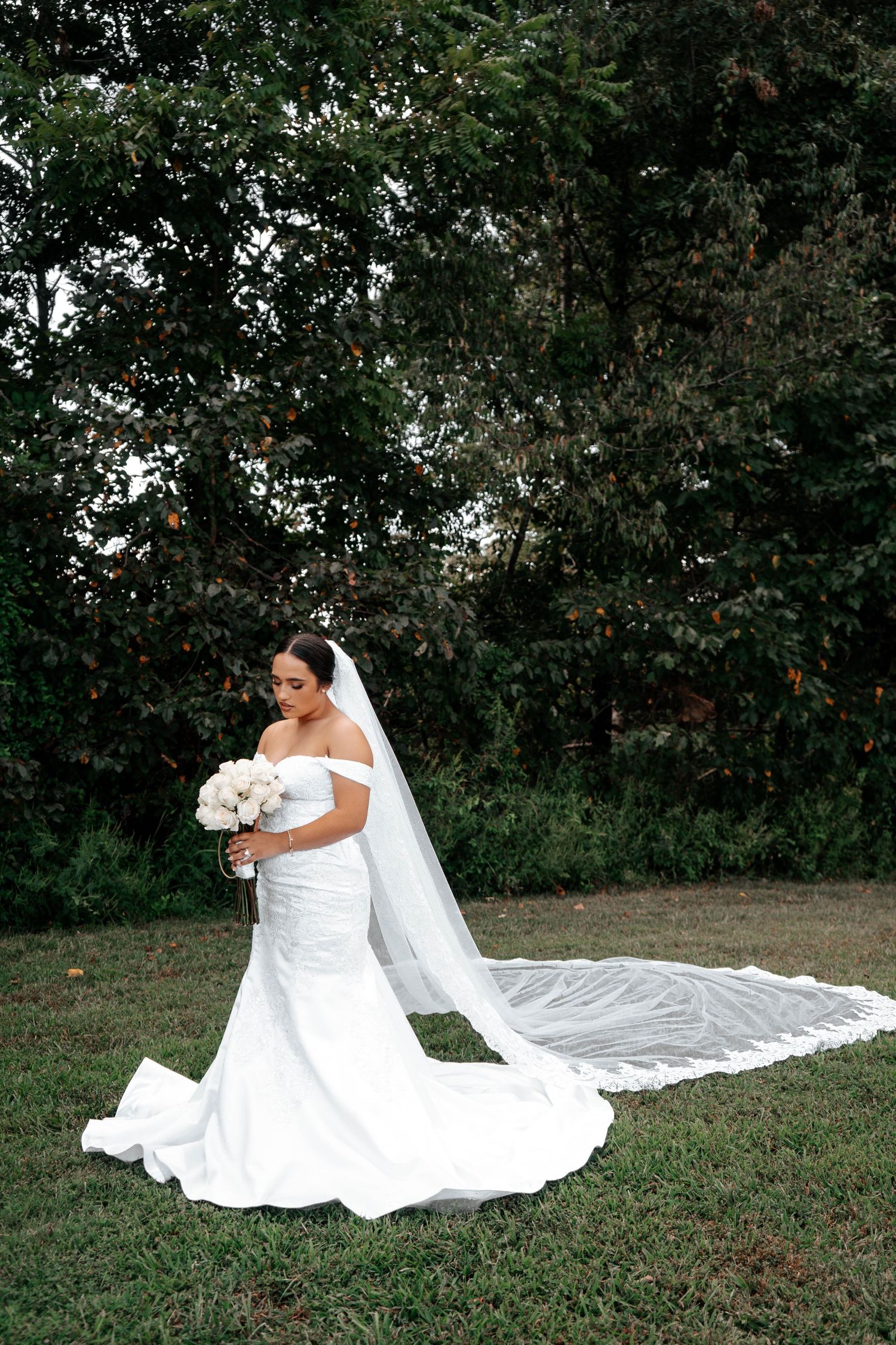 Bride in off-shoulder mermaid gown with cathedral veil holding white bouquet, standing on green lawn with trees behind.