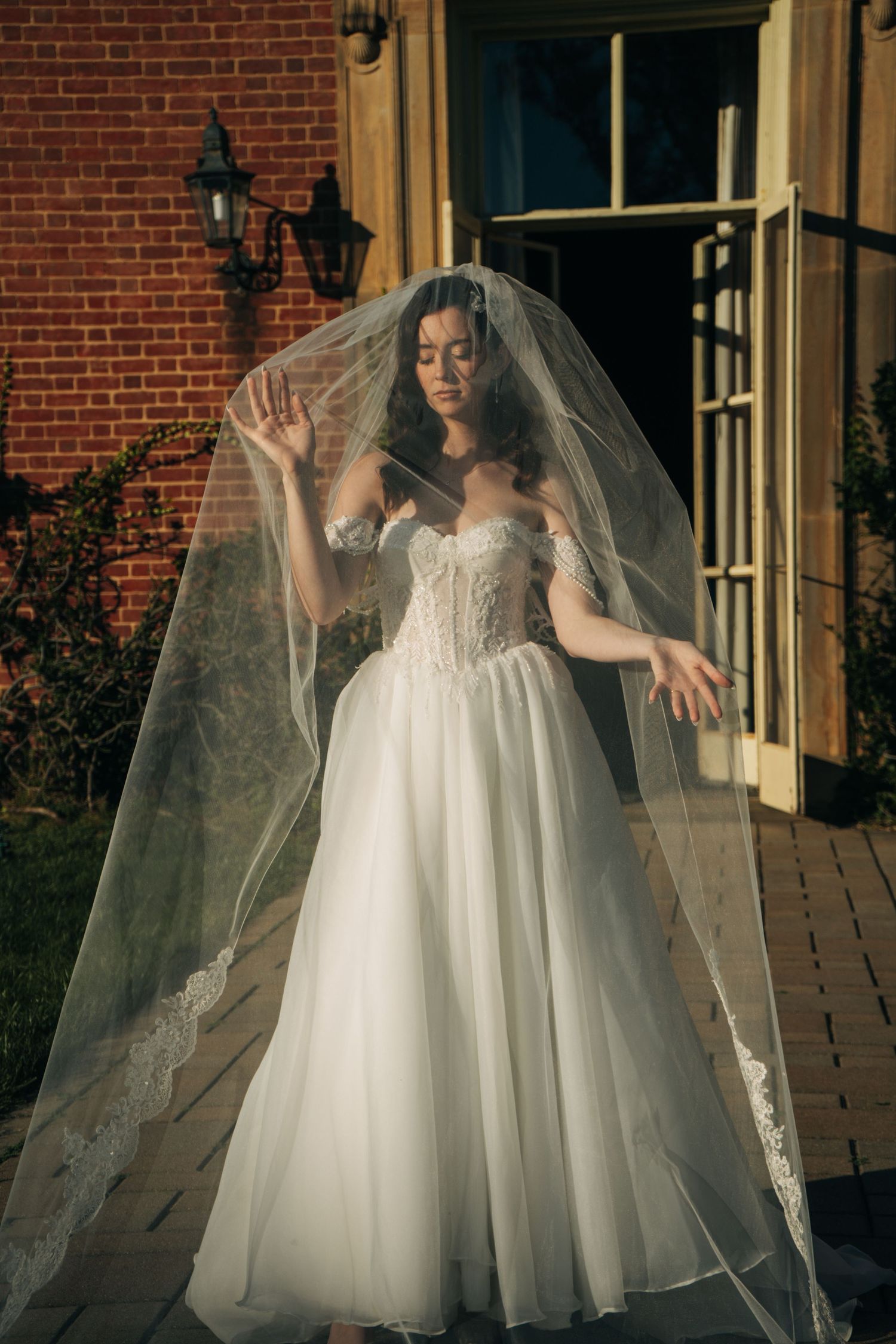 Bride in off-shoulder lace wedding gown with flowing veil poses gracefully outside brick building.