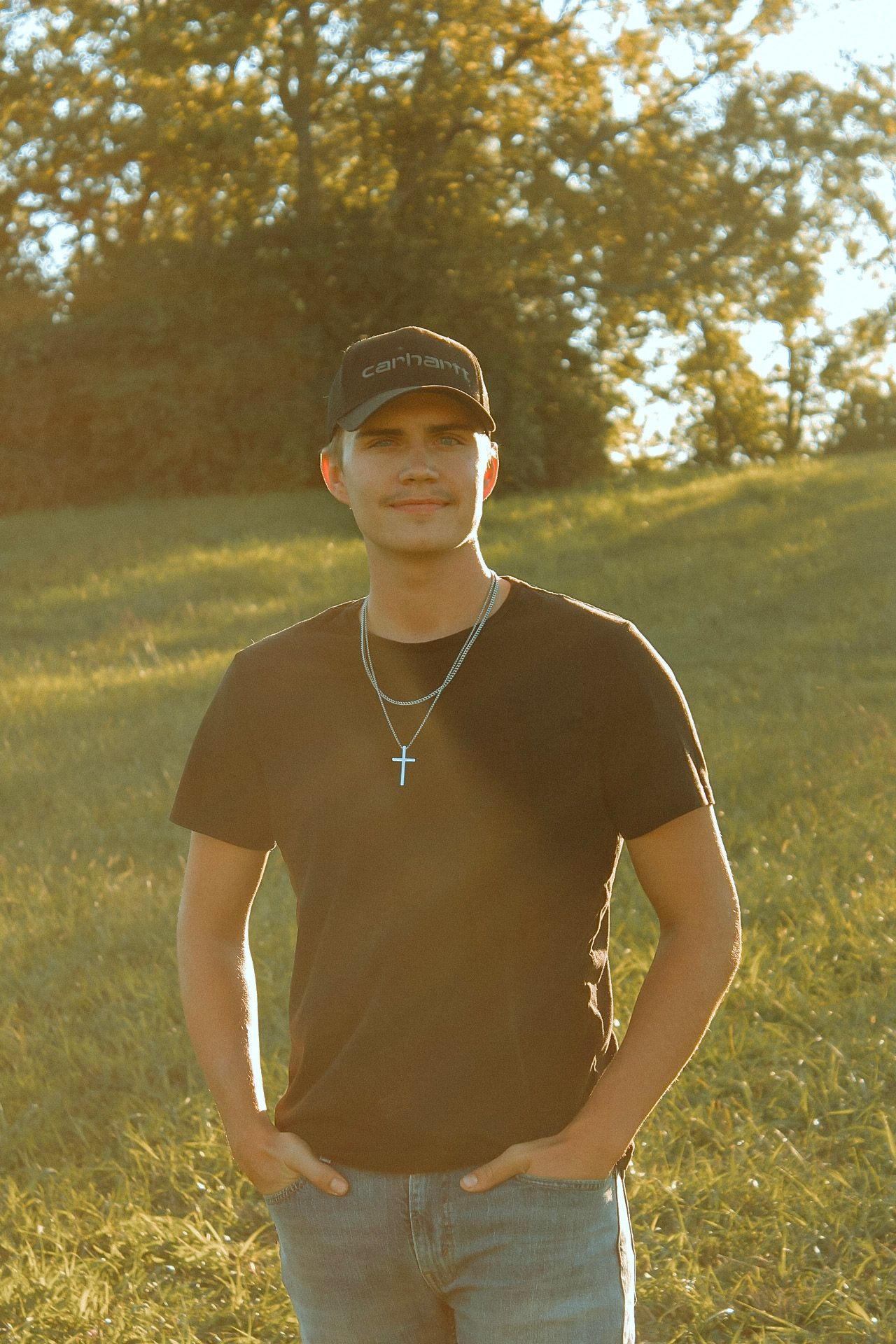 Young man in black cap and cross necklace poses in sunlit green field at golden hour.