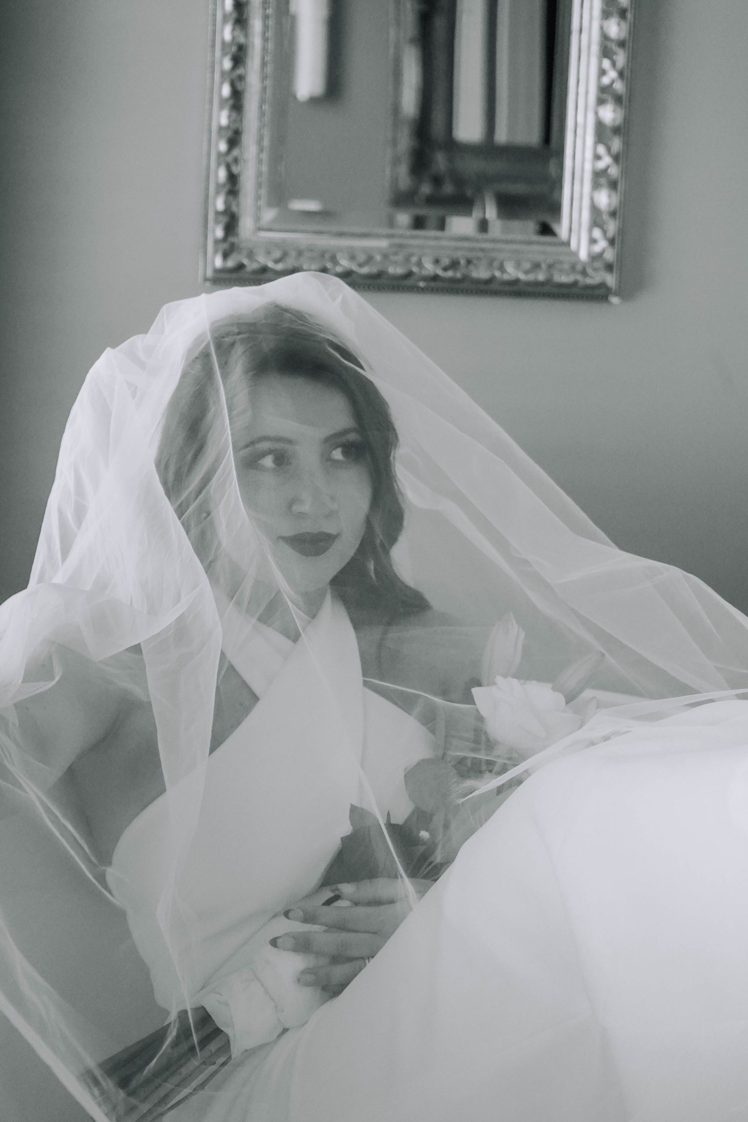 Black and white portrait of bride draped in sheer veil sitting in elegant room with ornate mirror.