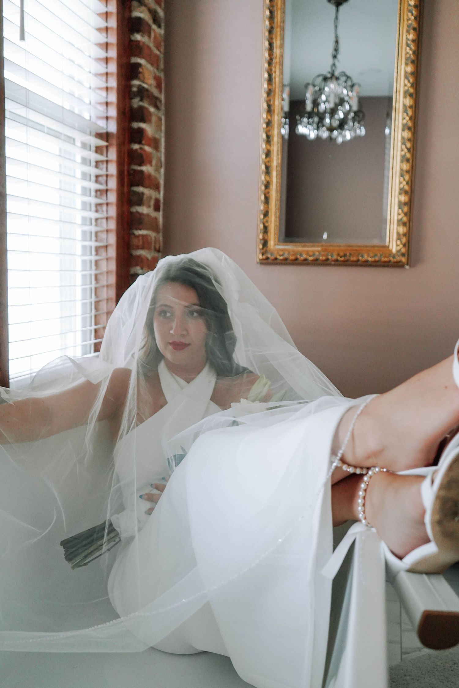 Bride in white gown and veil sitting near window with pearl heels reflecting in mirror behind her.