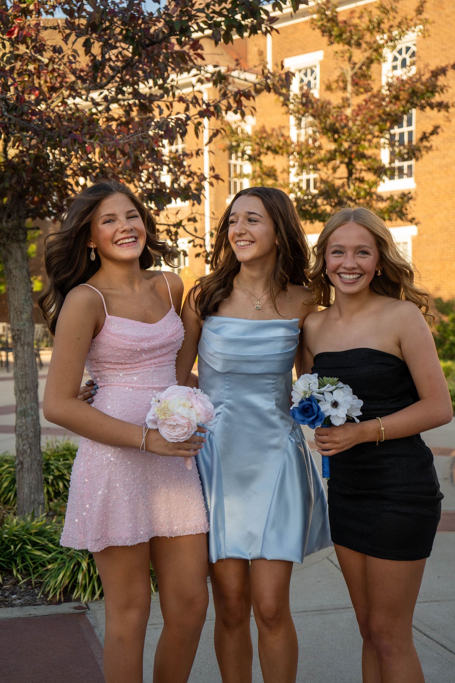 Three smiling teen girls in formal dresses pose together outdoors before a school dance.
