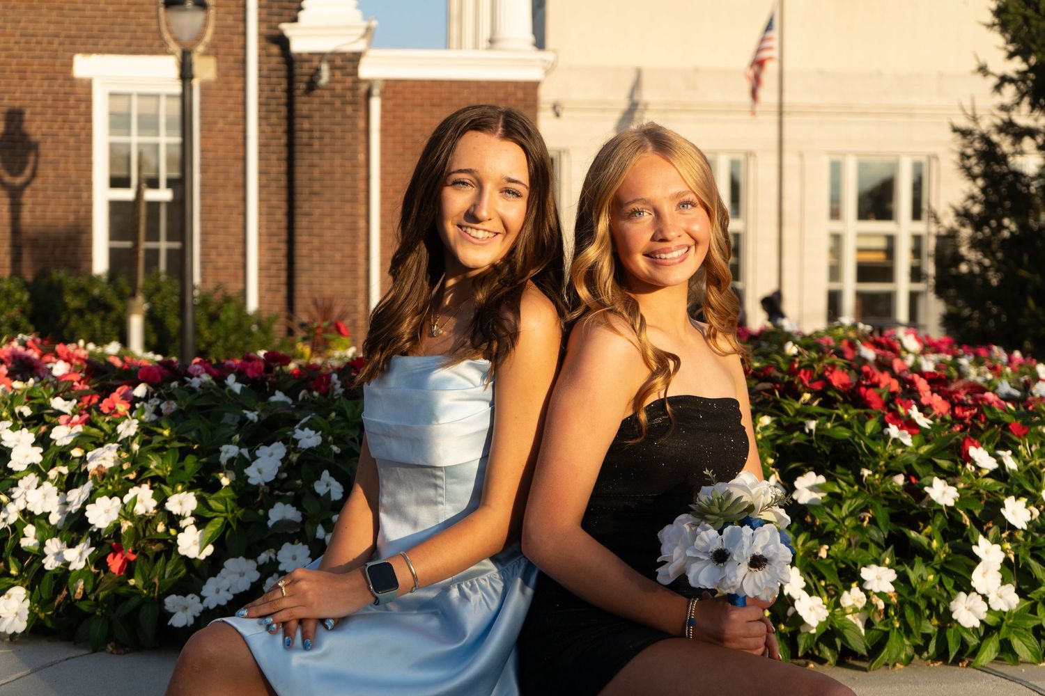 Two smiling teenage girls in blue and black formal dresses pose together by garden flowers.