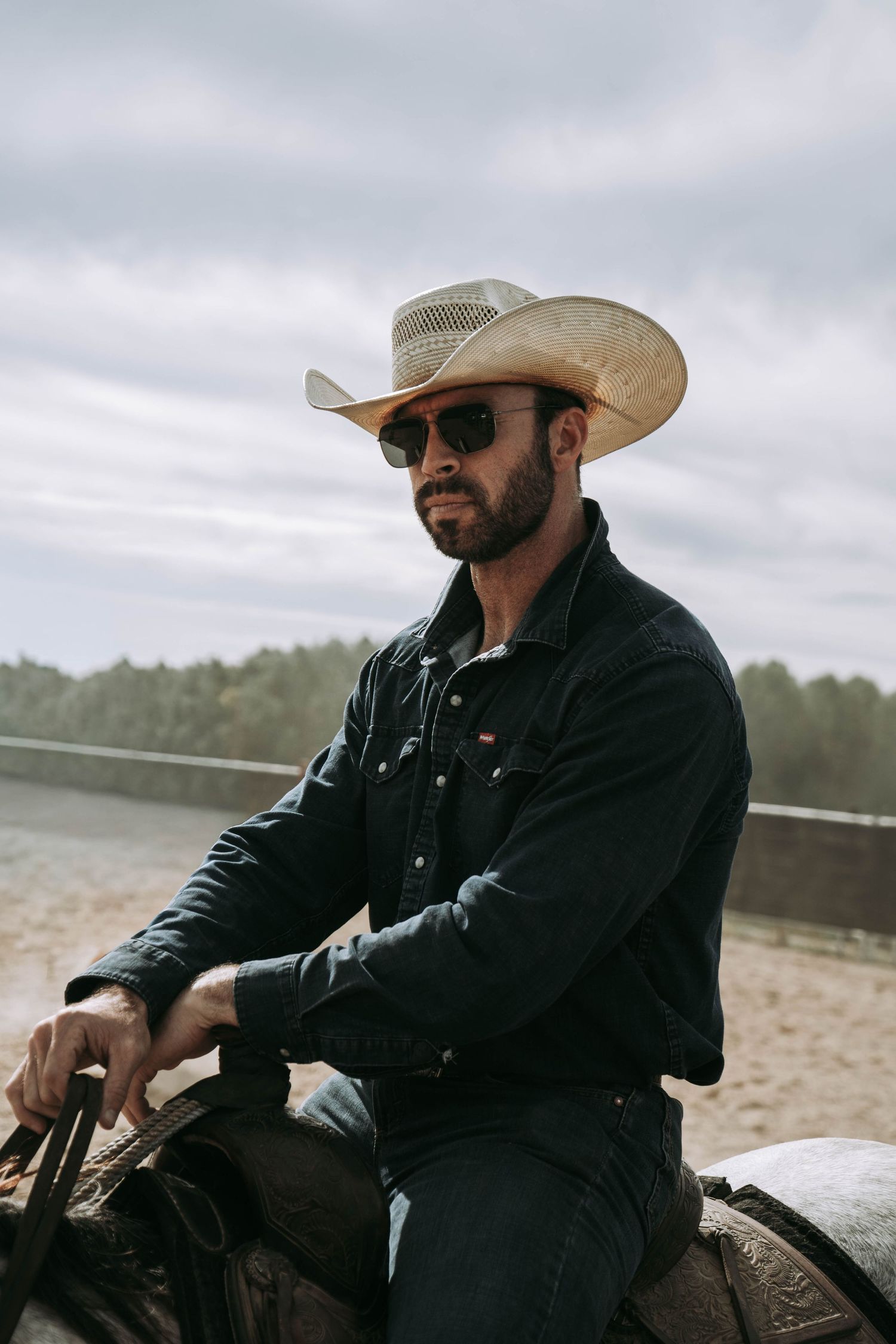 Bearded cowboy in straw hat and sunglasses sitting on horseback at outdoor sandy riding arena.