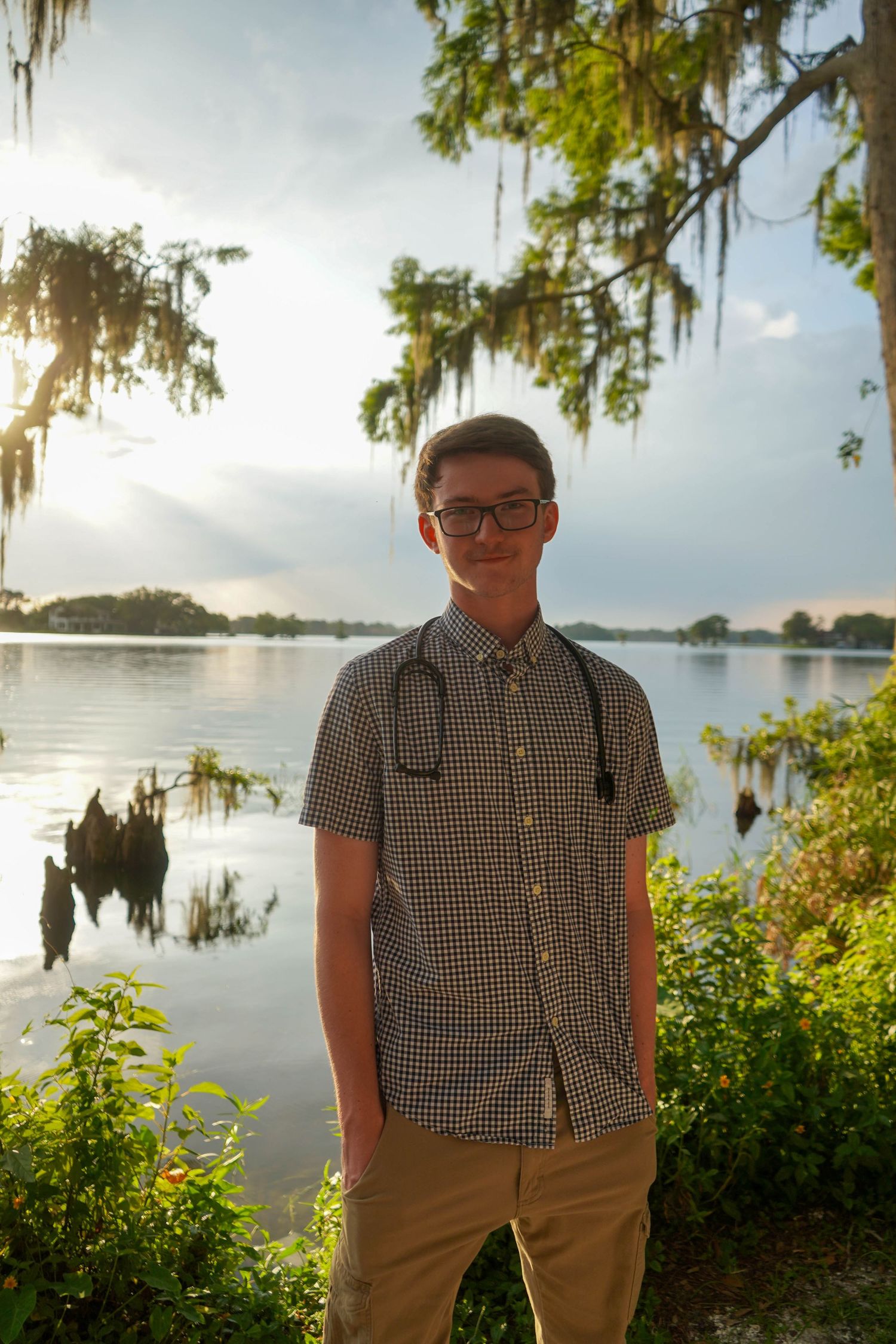 Young medical student with stethoscope standing by a serene lake surrounded by Spanish moss trees at sunset.