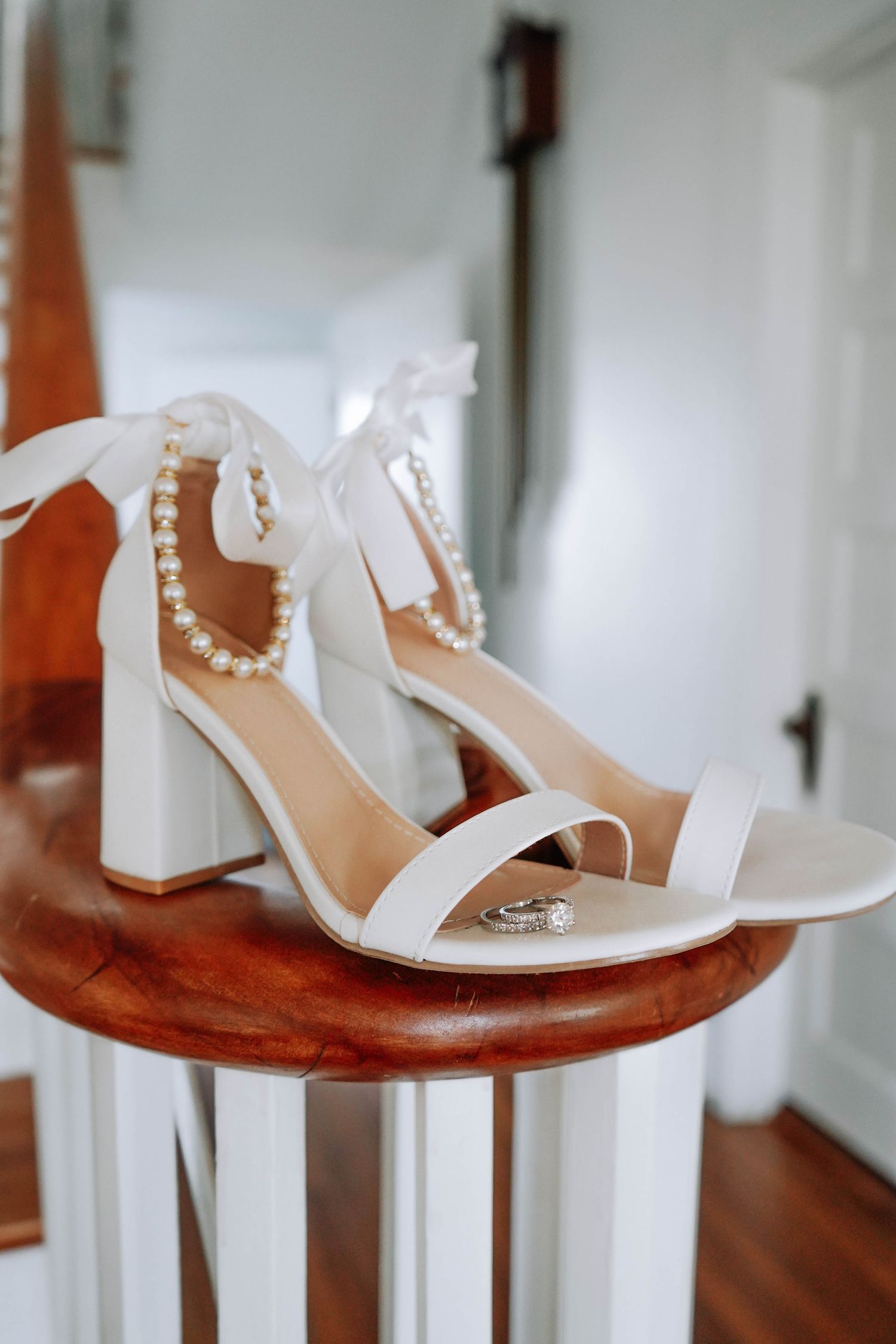 White pearl-strap block heel bridal sandals displayed on wooden banister with diamond wedding rings placed inside.