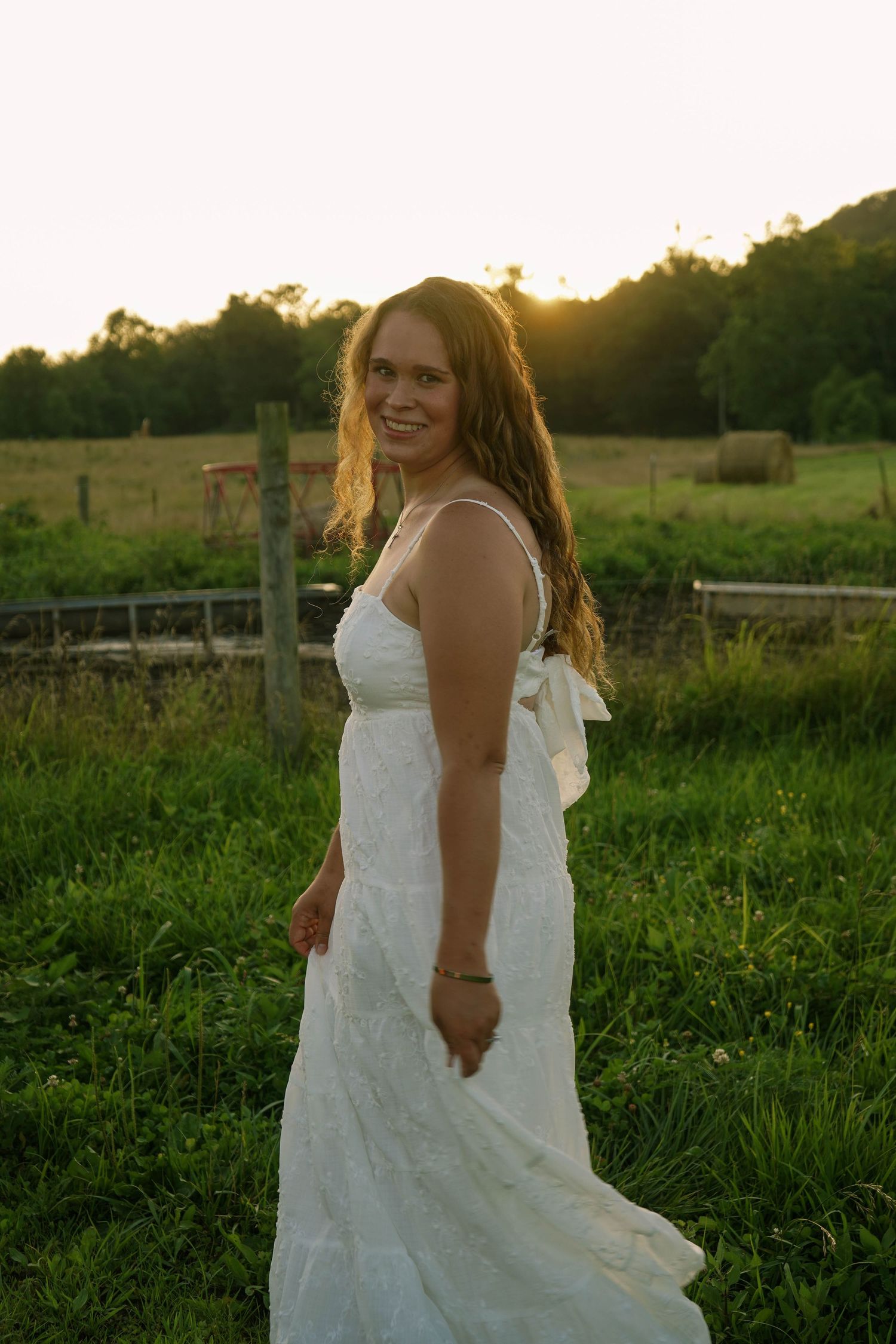 Young woman in white bow-back dress smiling in a lush green field at sunset.