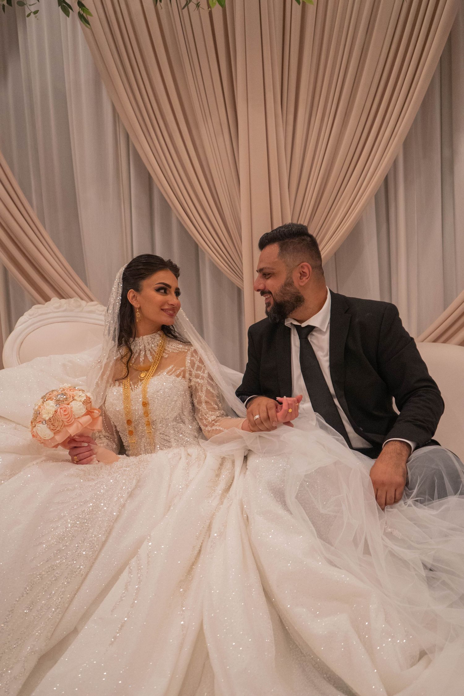 Bride in ornate gold-detailed gown and groom in black suit hold hands at wedding reception.
