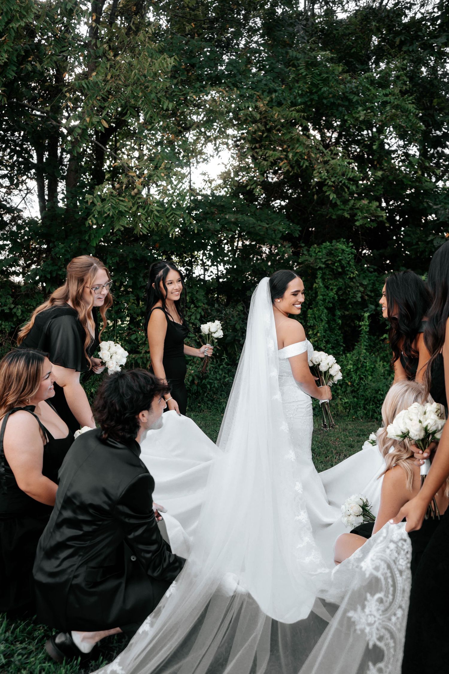 Bridesmaids in black dresses help bride with white lace veil outdoors surrounded by lush green trees.