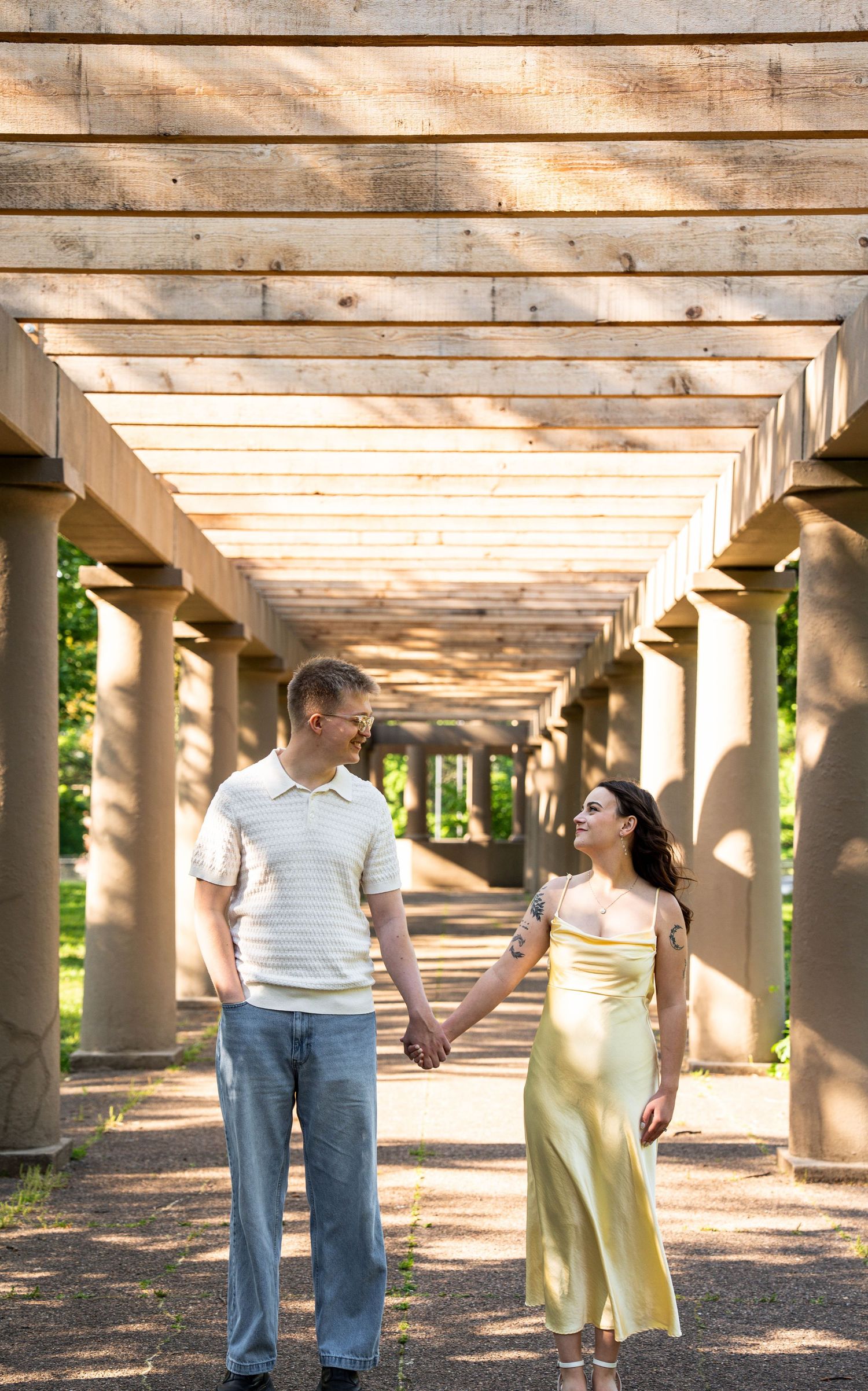Couple holding hands and smiling at each other under a wooden pergola in a park.