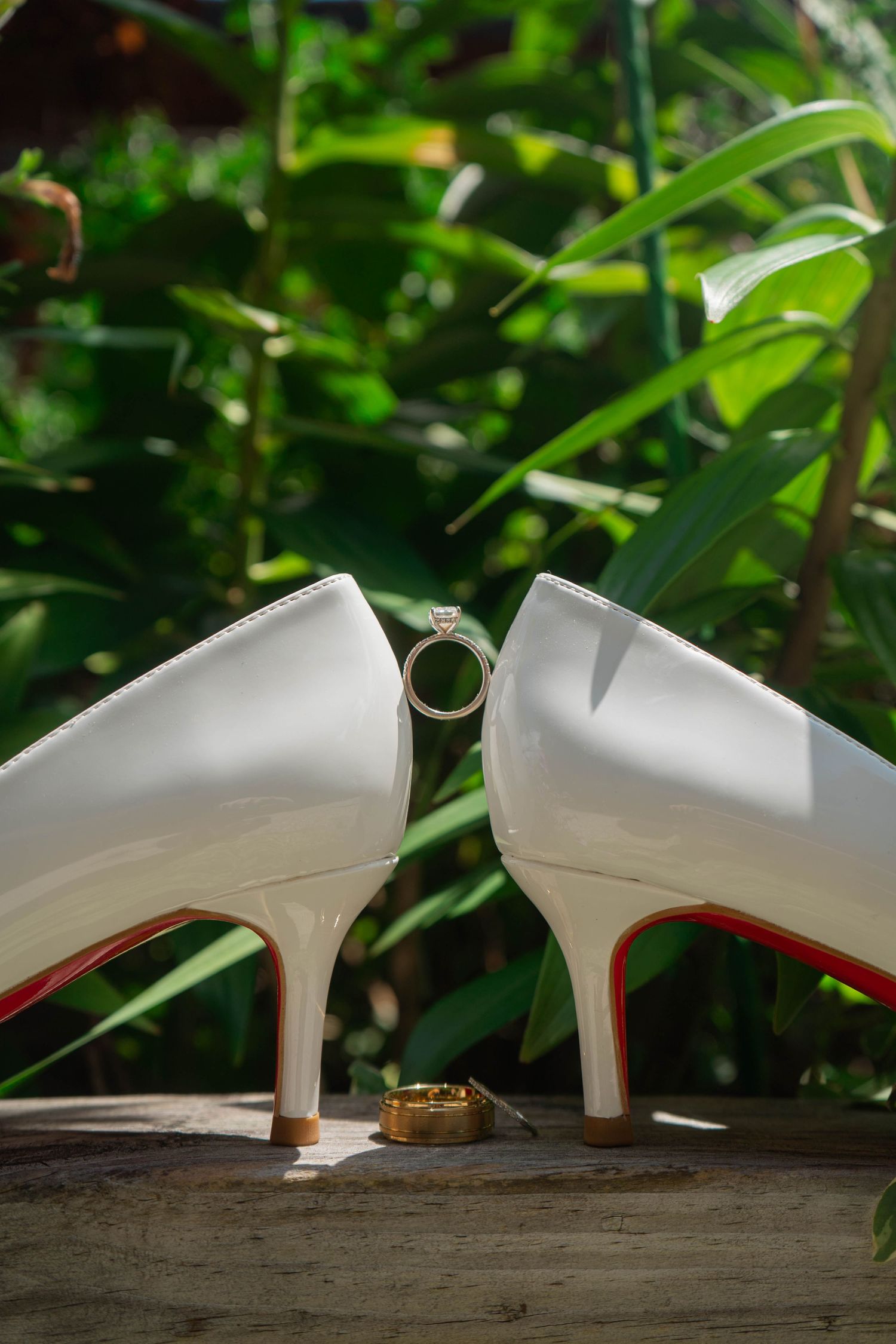 White Christian Louboutin bridal heels with diamond engagement ring balanced between shoes outdoors.