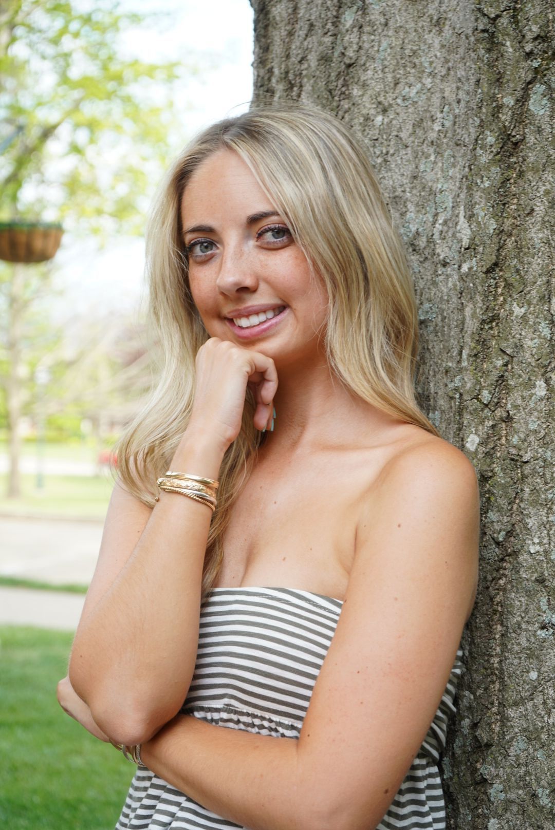 Blonde woman in striped strapless top leaning against tree trunk in sunny park.
