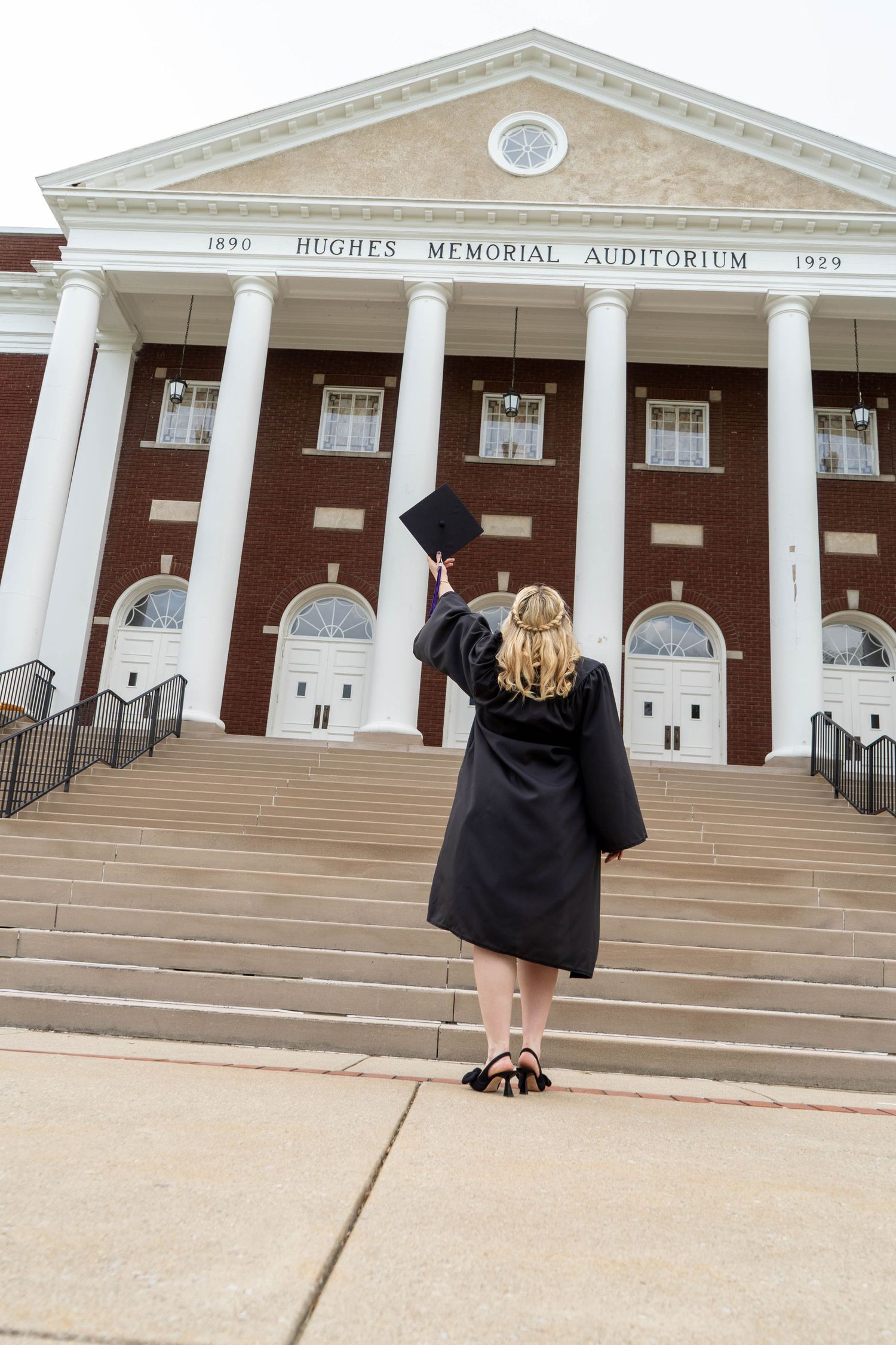 Graduate in black cap and gown raising mortarboard before Hughes Memorial Auditorium.