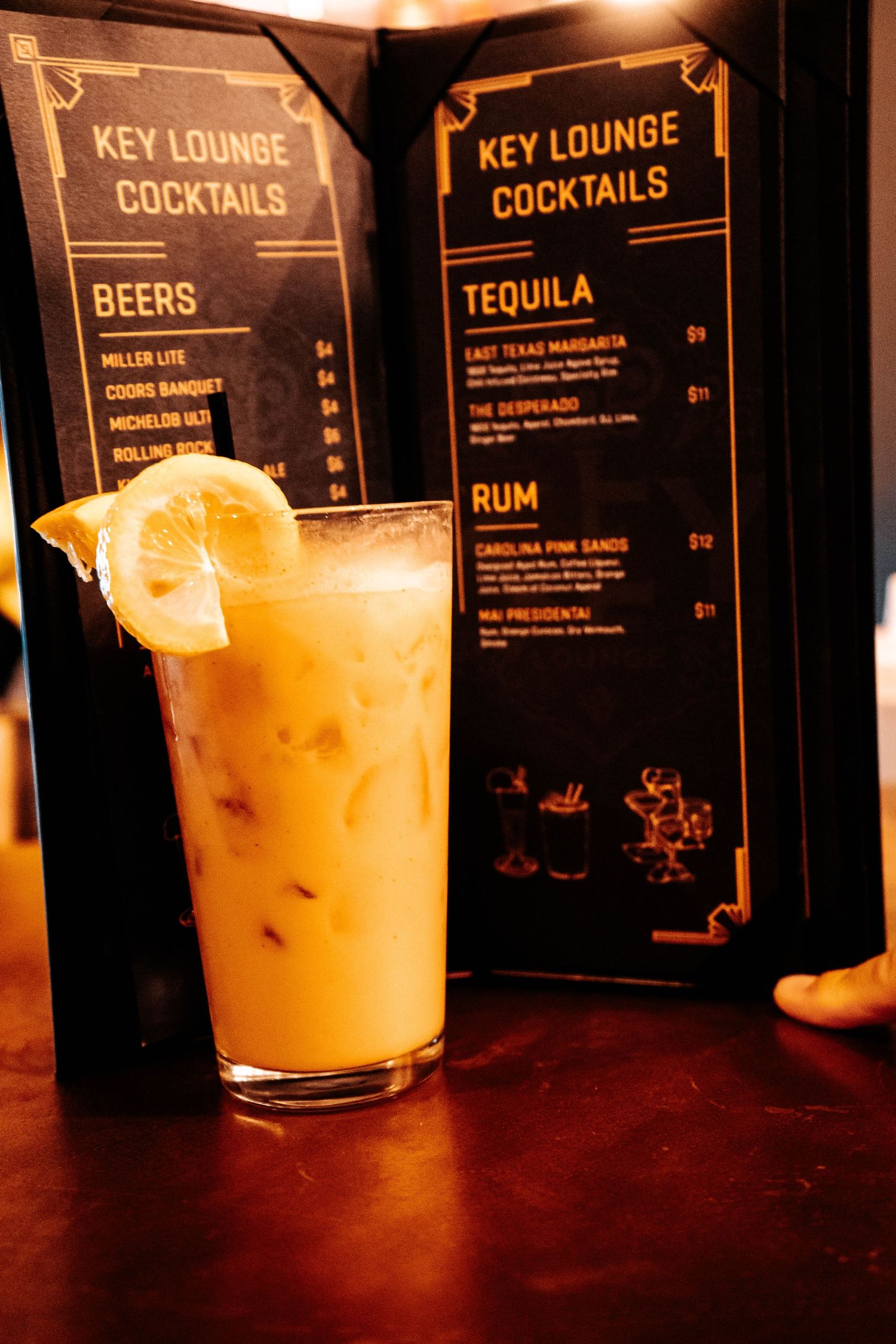 A tall cocktail with lemon garnish at Key Lounge bar, with cocktail menus featuring tequila and rum drinks in background.