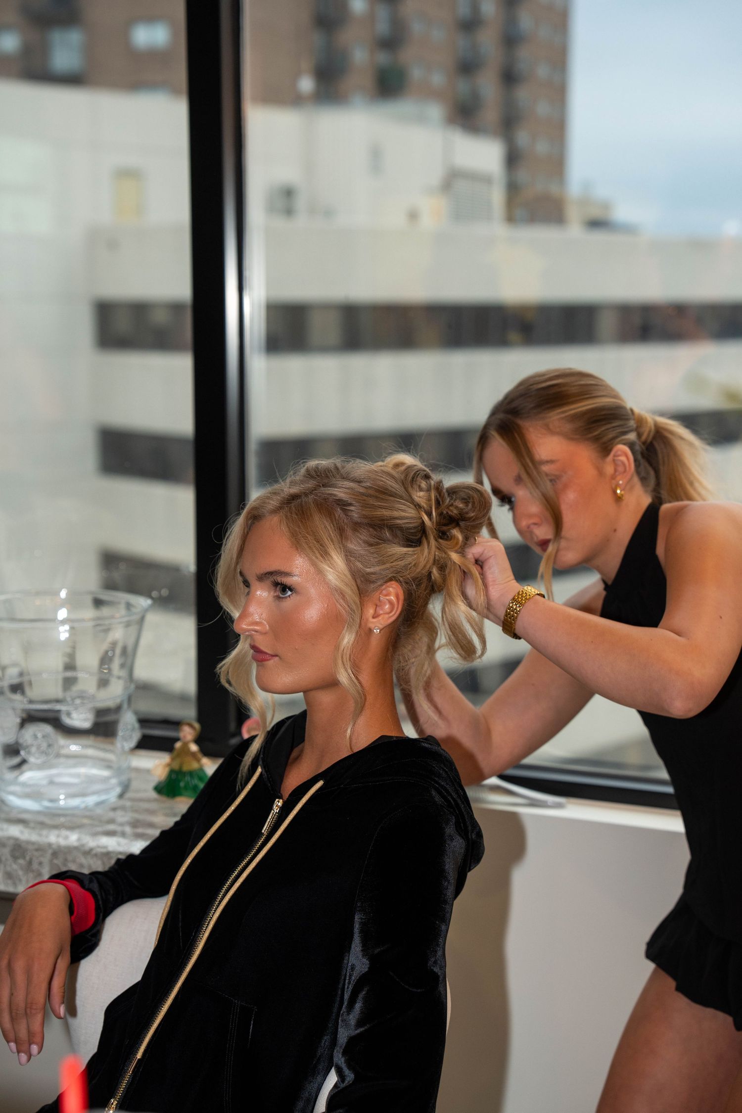 Hairstylist styling blonde woman's updo in black velvet dress, city skyline visible through window.