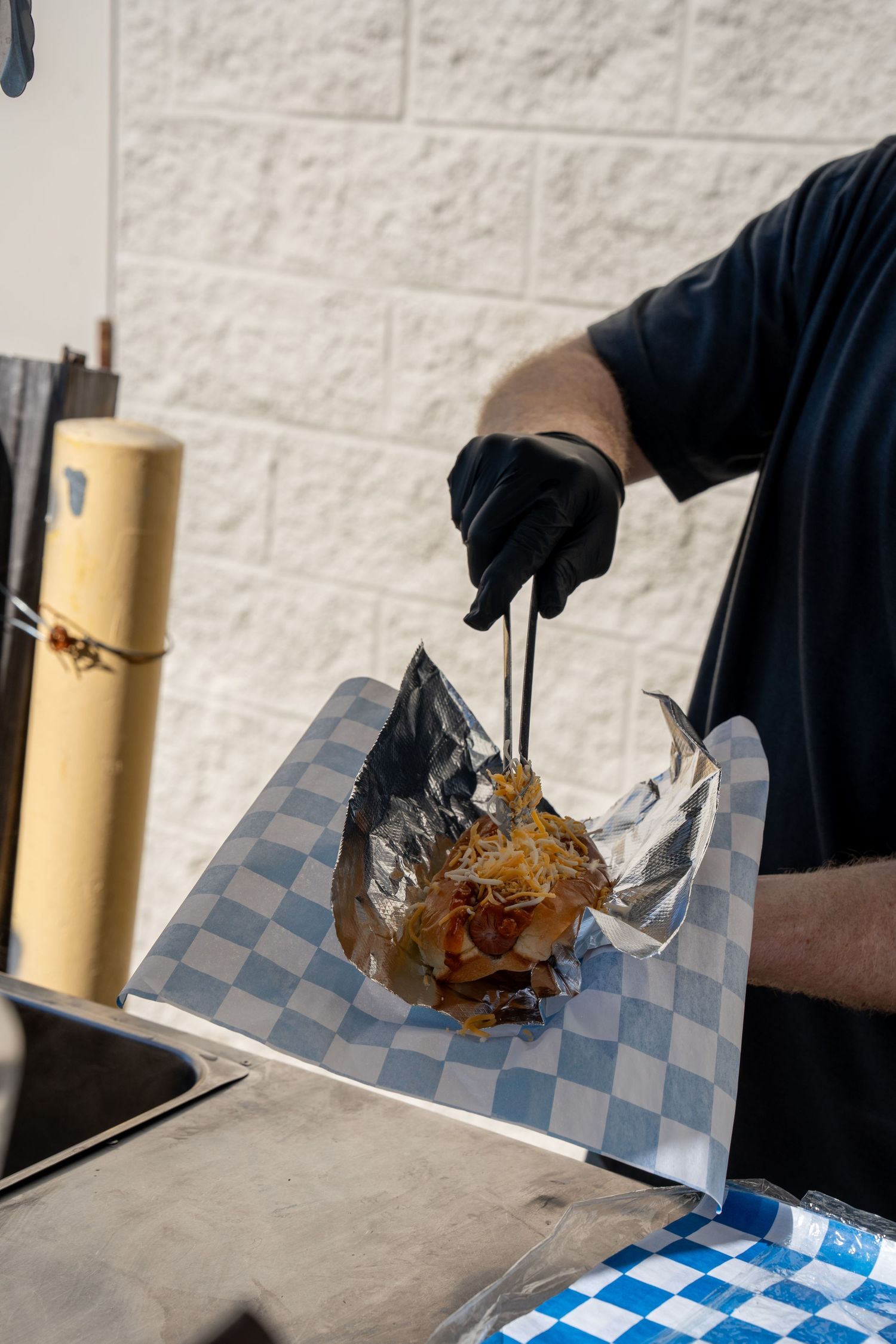 Chef in black gloves uses tongs to plate a loaded street food dish on checkered deli paper at an outdoor food stand.