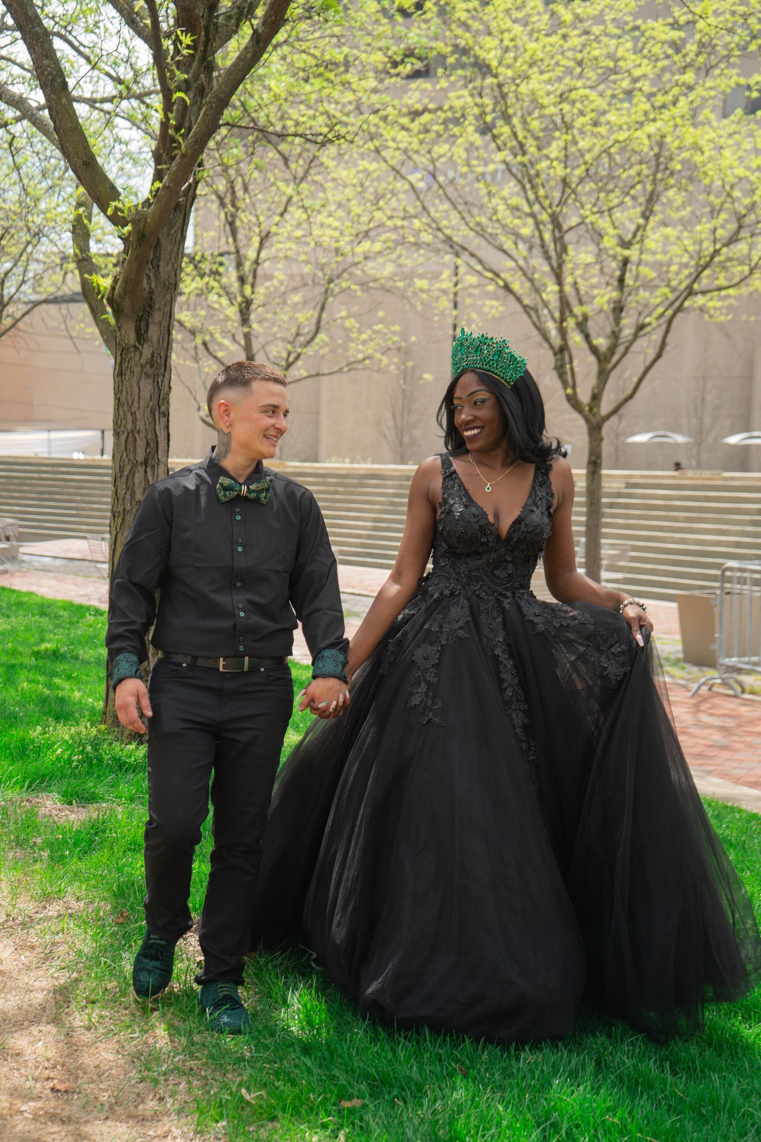 Couple in matching black formal wear holding hands outdoors, woman wearing black ballgown and green crown.