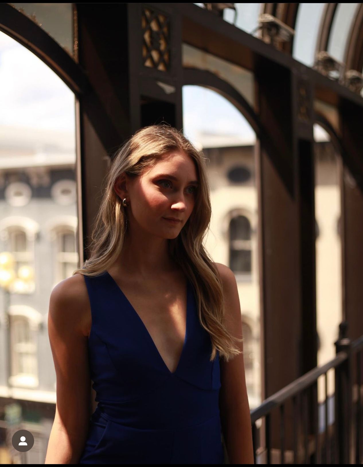 Elegant blonde woman in royal blue v-neck dress stands near ornate iron balcony railing.