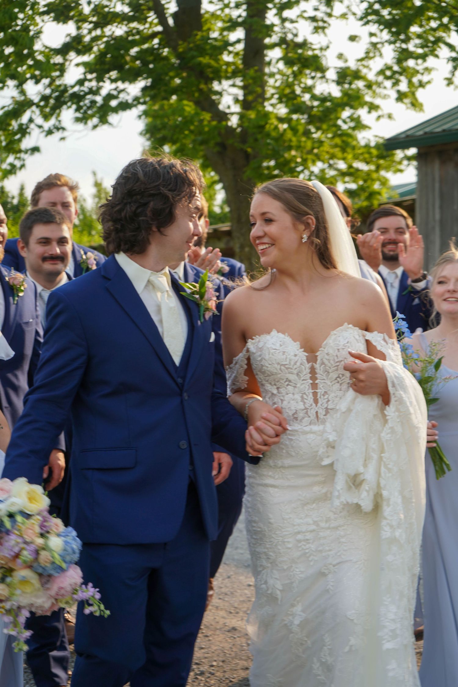Newlyweds walk joyfully through cheering guests, bride in lace gown and groom in navy suit after ceremony.