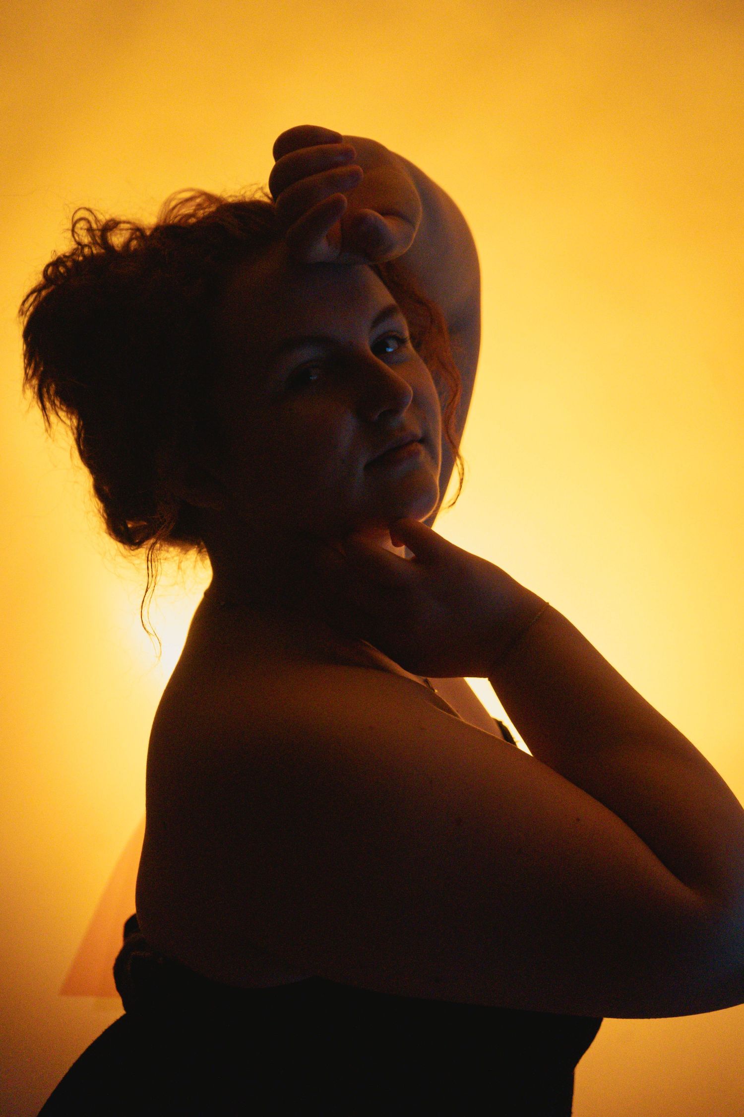 Dramatic silhouette of young woman posing with hand raised against vibrant orange-yellow background.