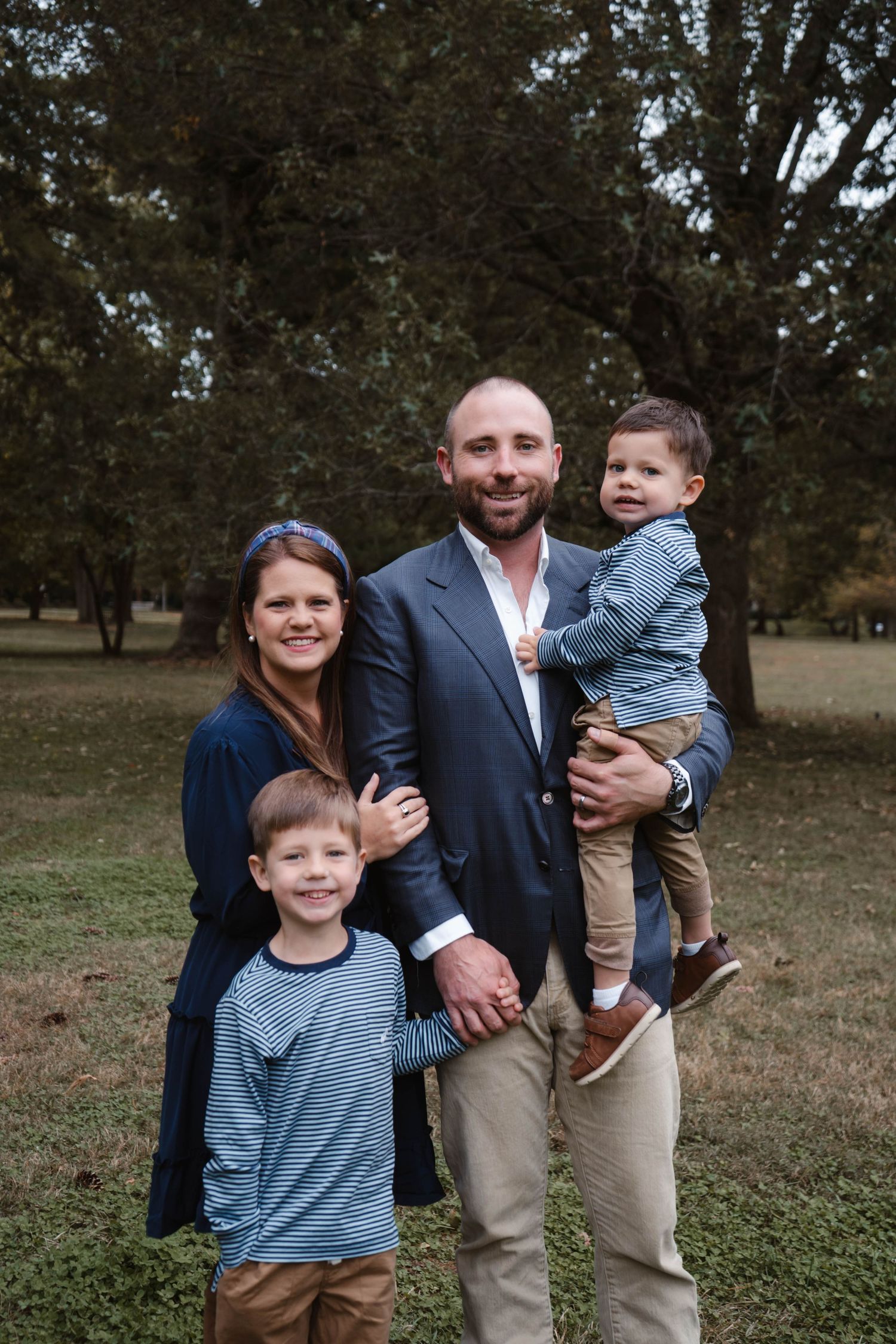Happy family of four poses outdoors, father holding toddler while mother and older son stand beside him in park.