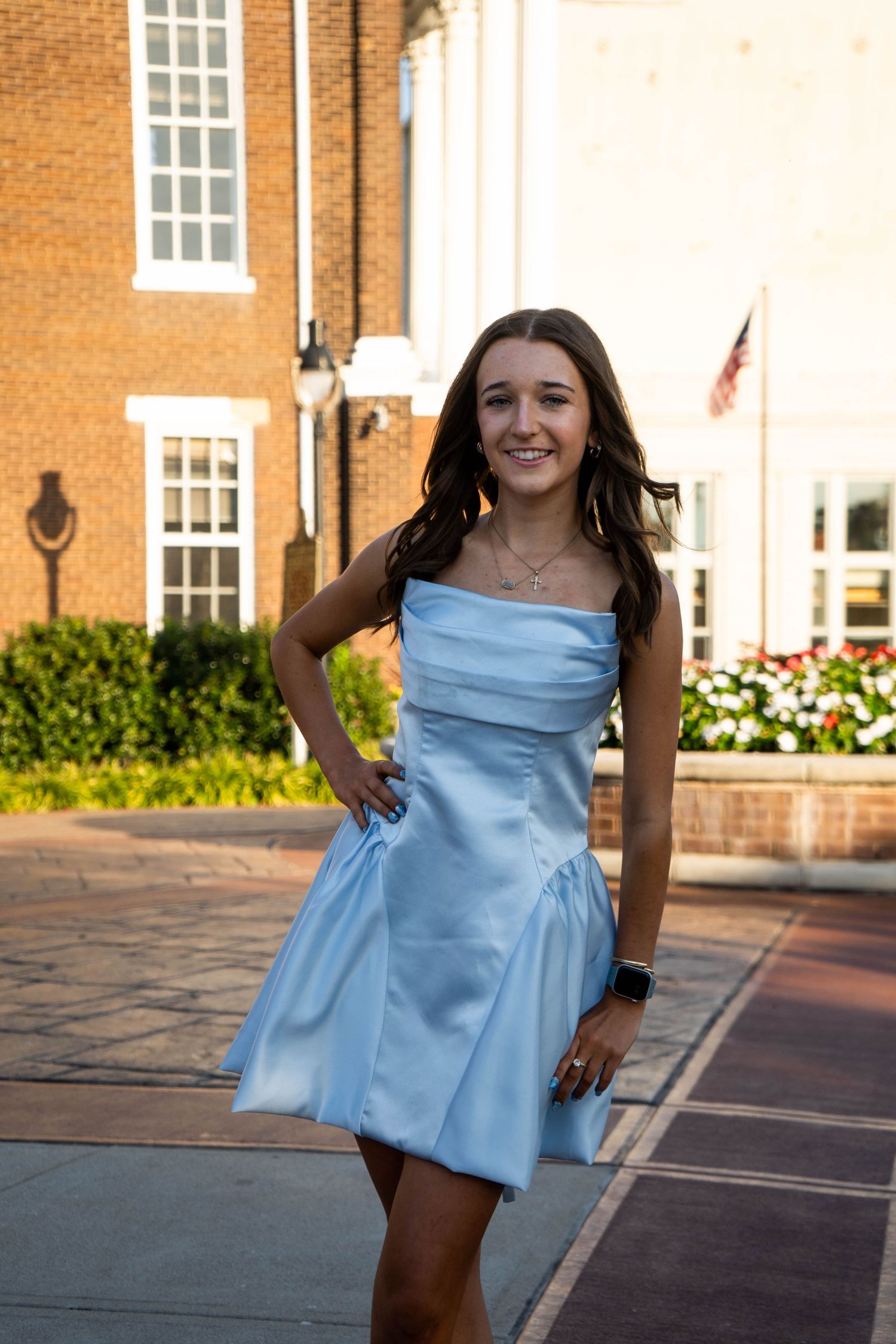 Young woman in powder blue strapless dress poses gracefully outdoors near brick building at sunset.